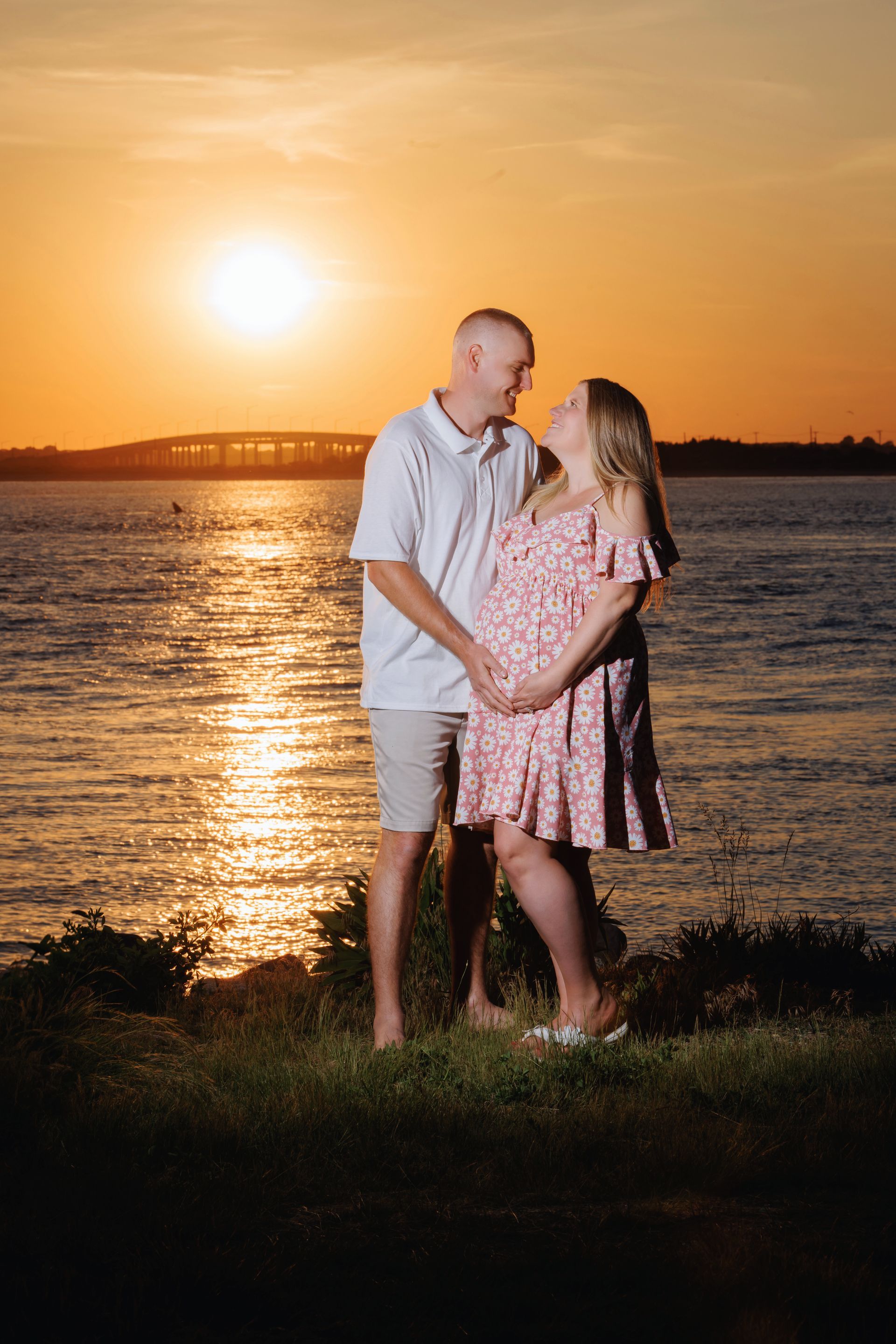 Couple embracing on a grassy shore at sunset with golden water and sky behind them