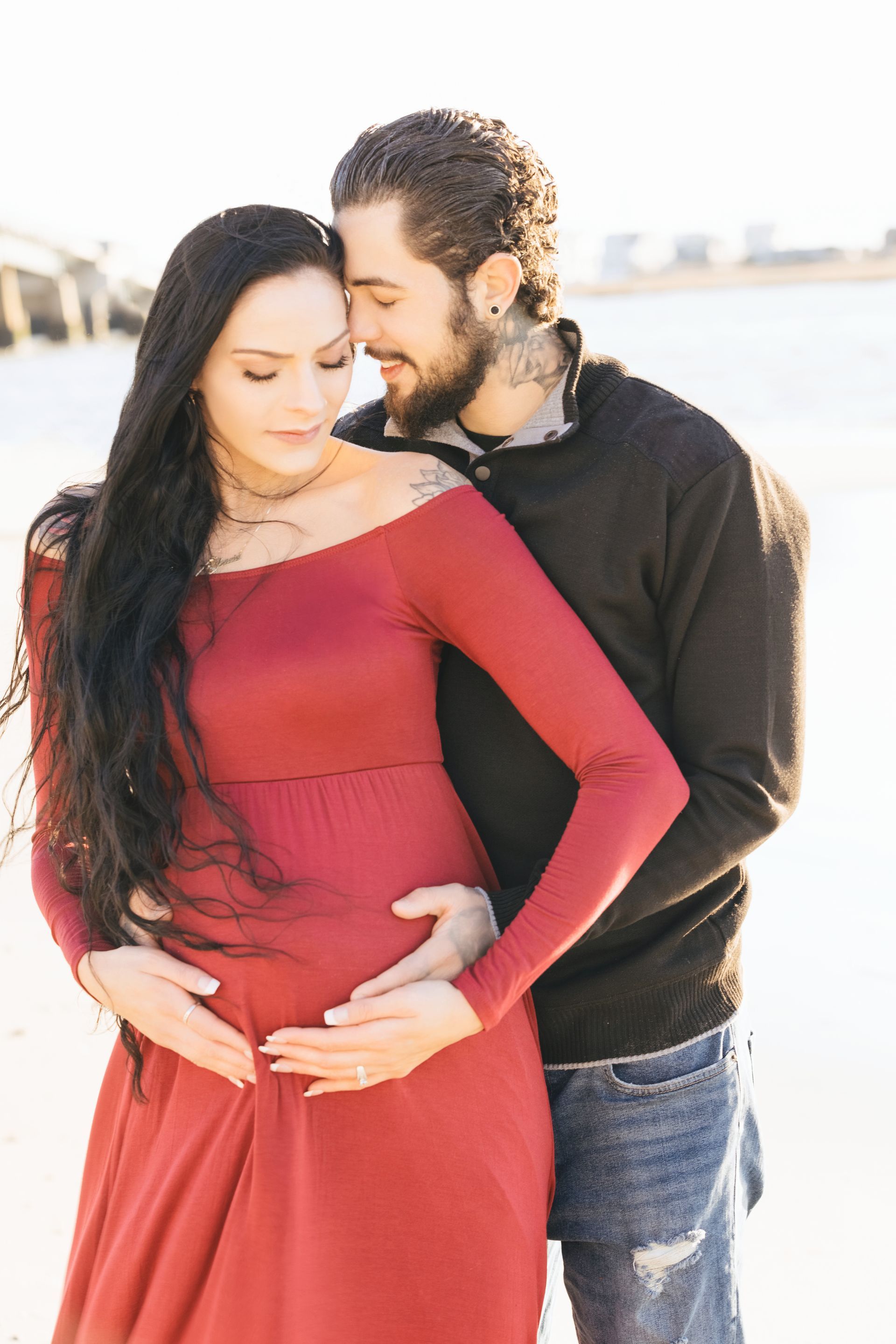 Couple standing outdoors, gently holding a pregnant belly, with a bright beach-like background.