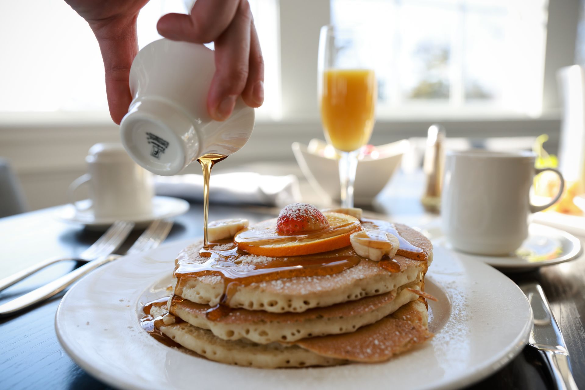 Syrup being poured over a stack of pancakes on a white plate at a bright breakfast table
