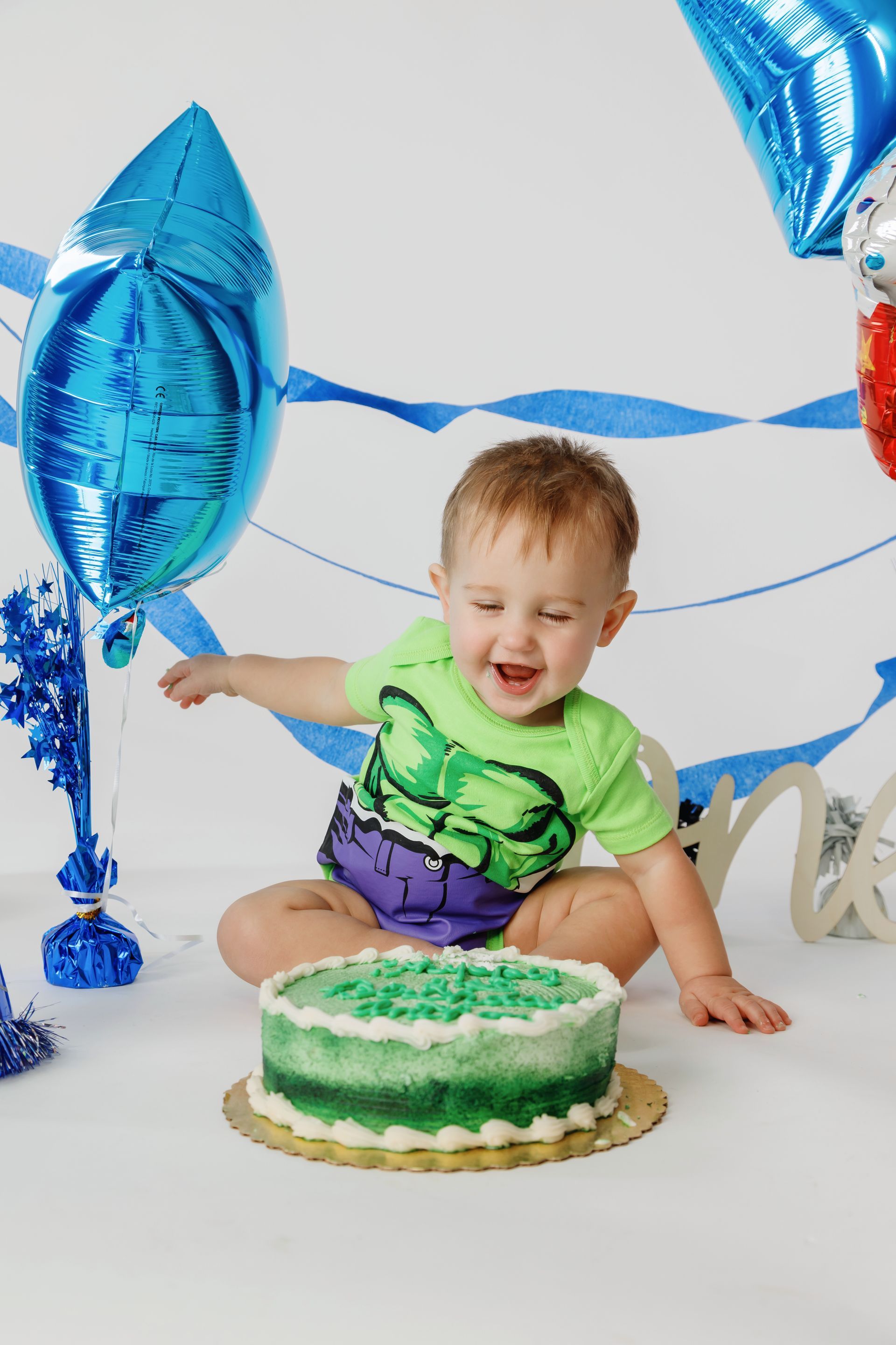 Smiling toddler in green shirt and purple shorts reaching toward a green cake with blue balloons behind.