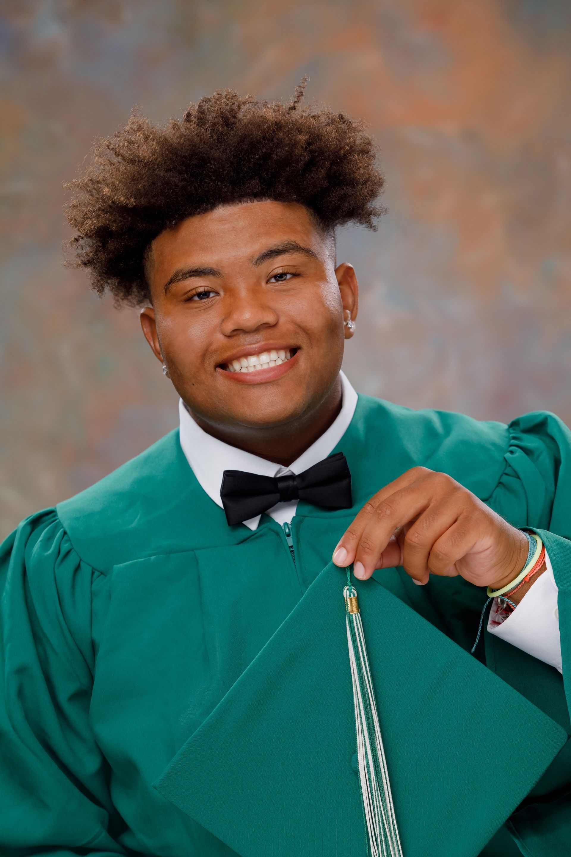 Graduate in green cap and gown smiling and holding a tassel against a studio backdrop