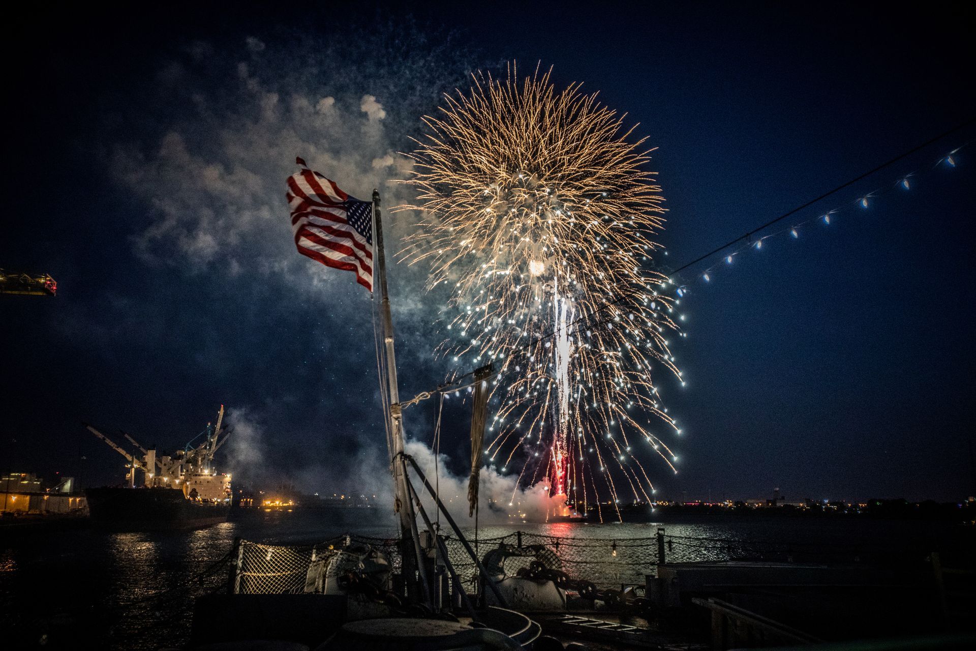 Fireworks over a waterfront at night with an American flag in the foreground