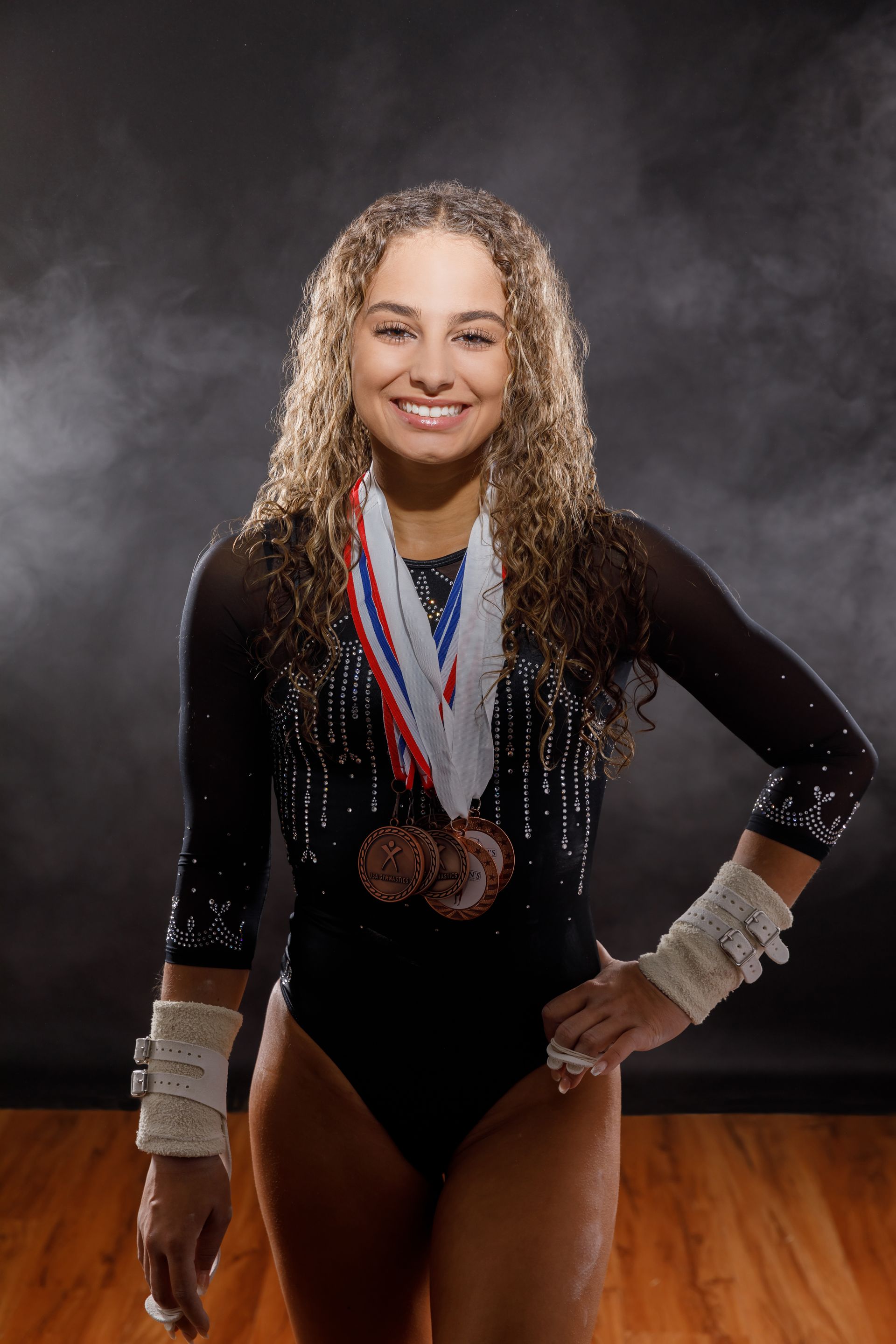 Gymnast posing in a black leotard with medals around her neck, standing on a gym floor.