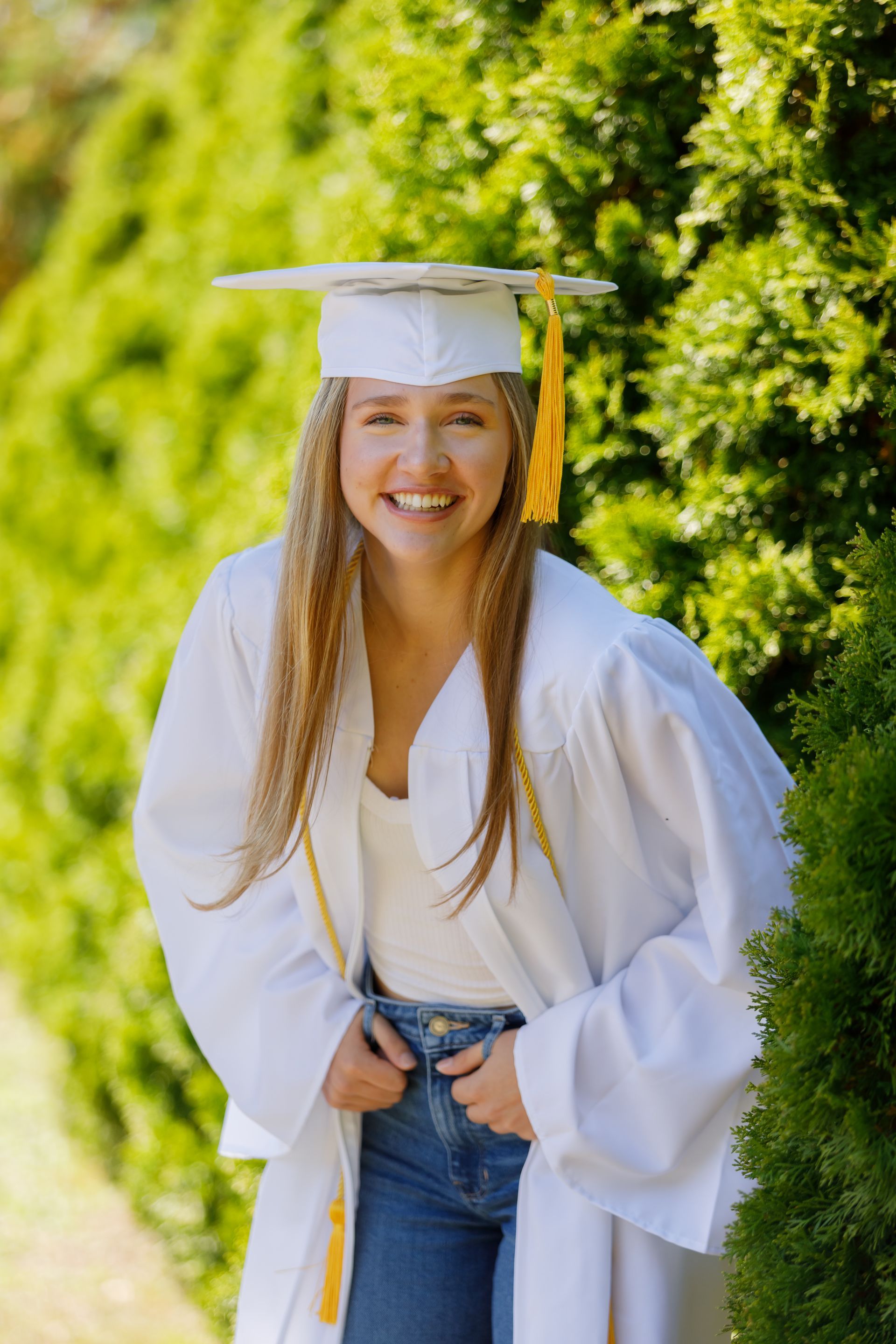 Smiling graduate in white cap and gown standing outdoors in a green garden