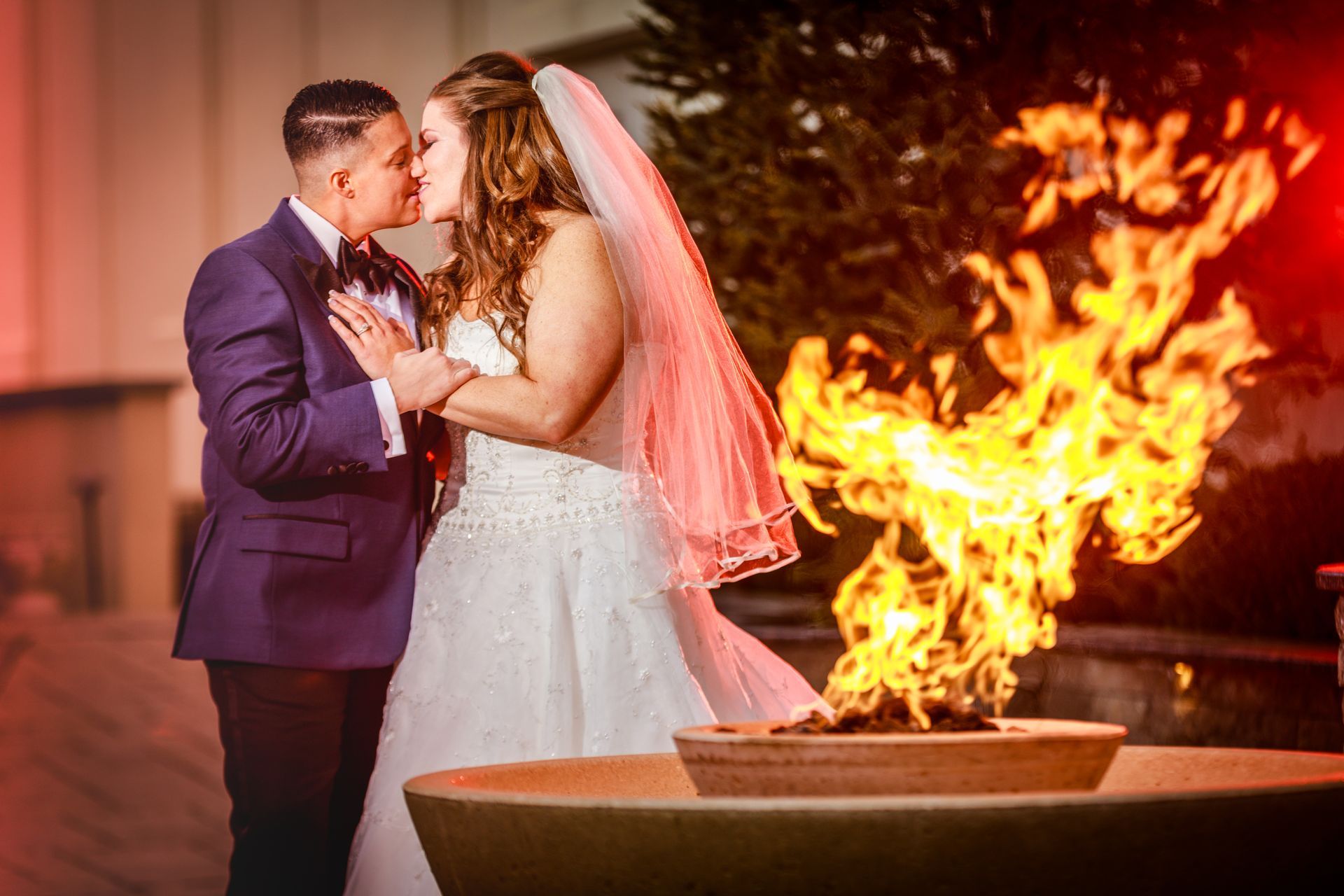 Bride and groom kiss beside a large fire pit at an outdoor wedding reception