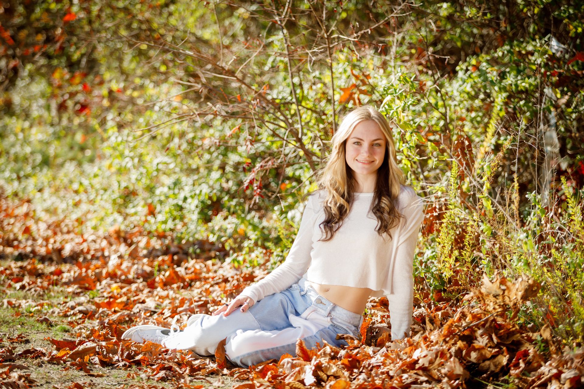 Woman sitting in autumn leaves beside a tree, wearing a white top and jeans, in a sunlit forest.