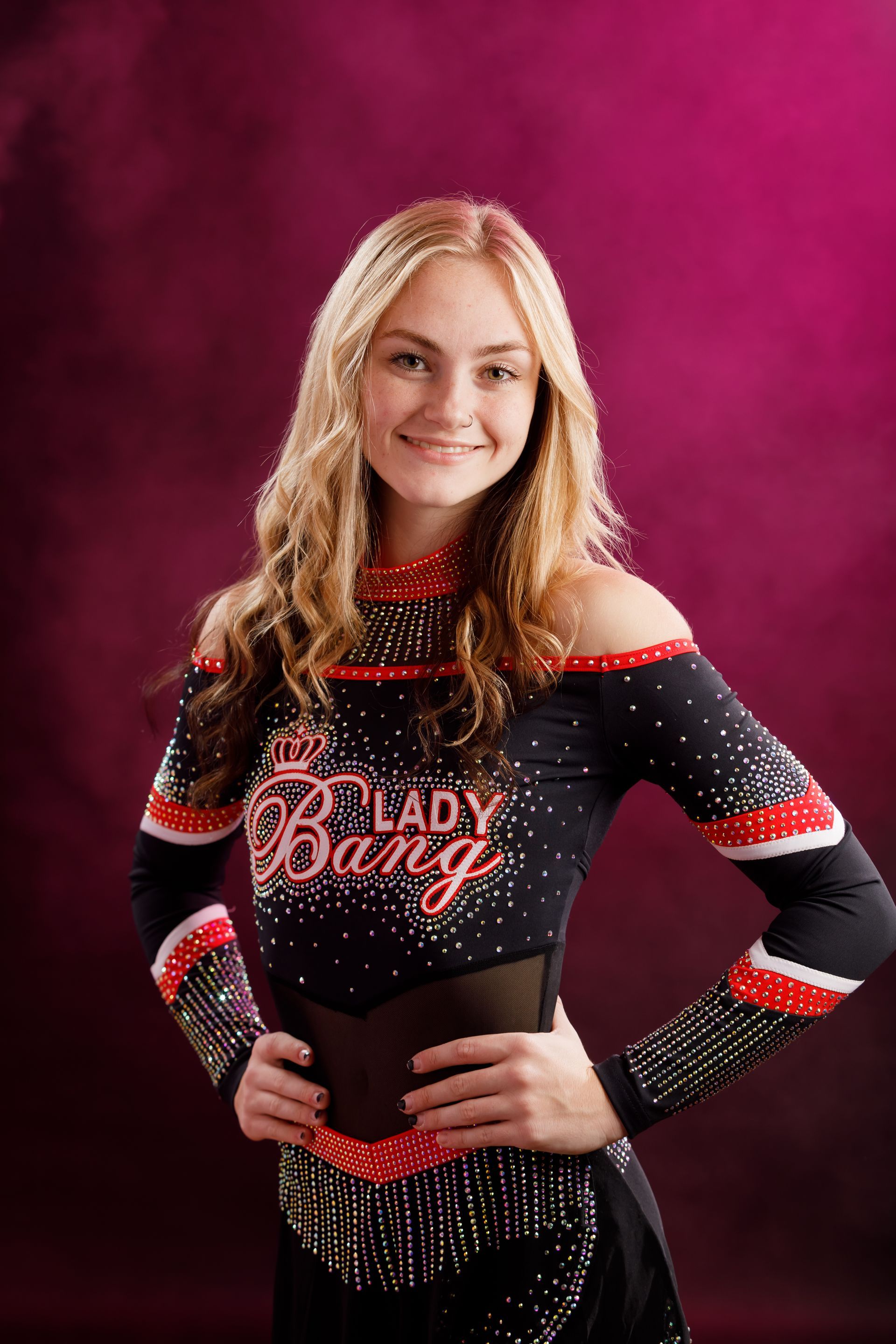 Smiling cheerleader in a black, red, and white outfit with fringe, standing hands on hips against a pink backdrop