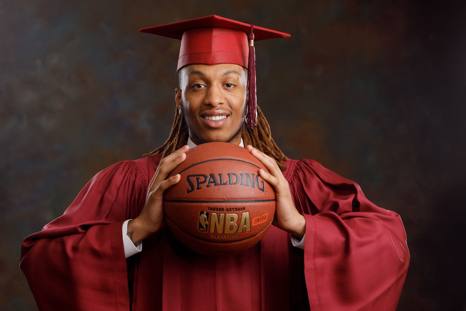 Graduate in red cap and gown holding a basketball against a dark studio background