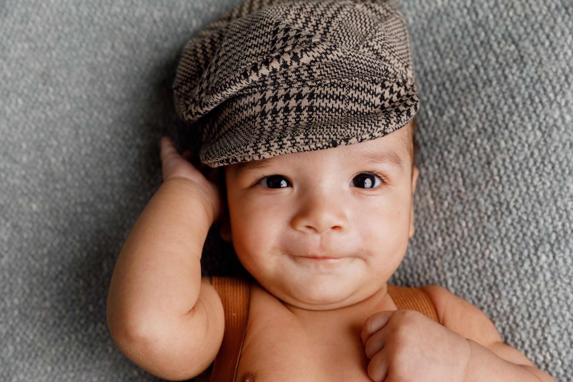 Infant lying on a gray blanket, wearing a patterned cap and touching their head.