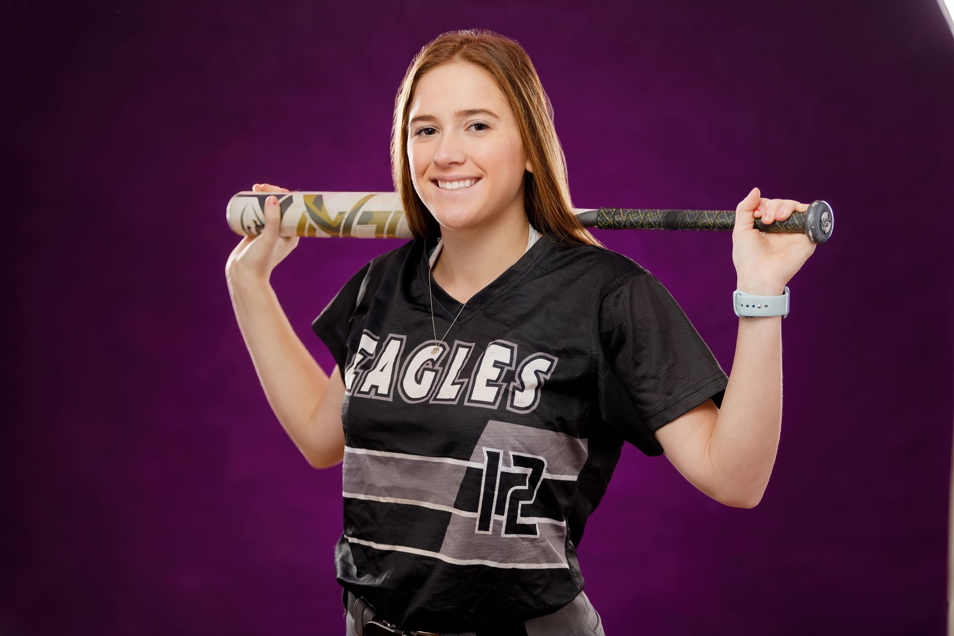 Smiling athlete in black jersey holding a baseball bat on her shoulders against a purple background