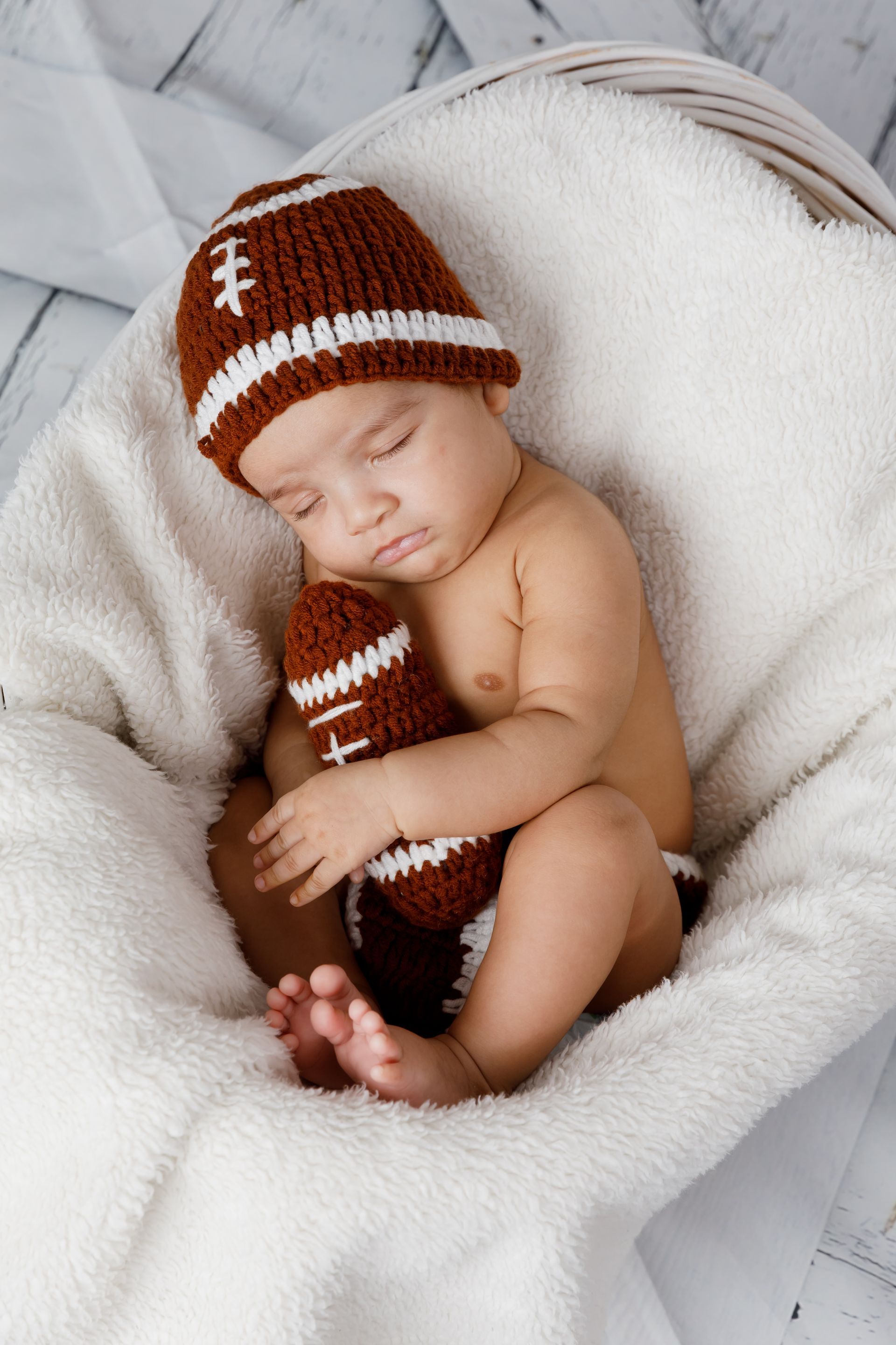 Sleeping baby in a brown football hat and scarf, curled on a white plush blanket