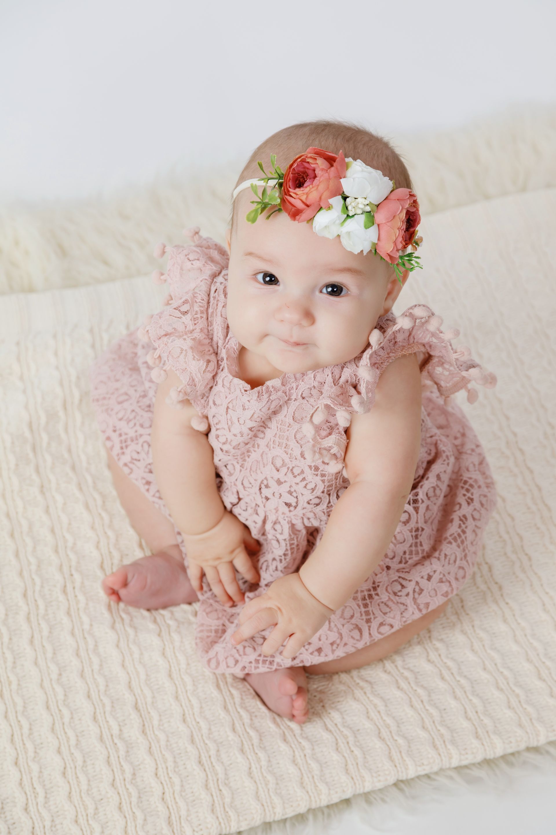 Baby in a pink dress and floral headband sitting on a white rug, smiling at the camera