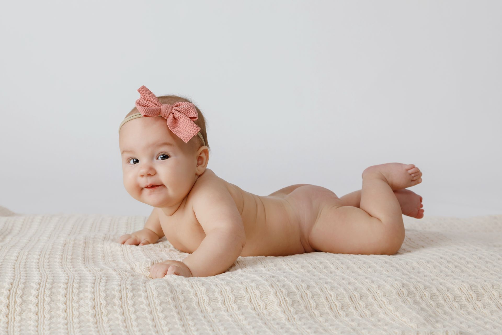 Baby lying on a cream blanket, wearing a pink bow headband and smiling at the camera.