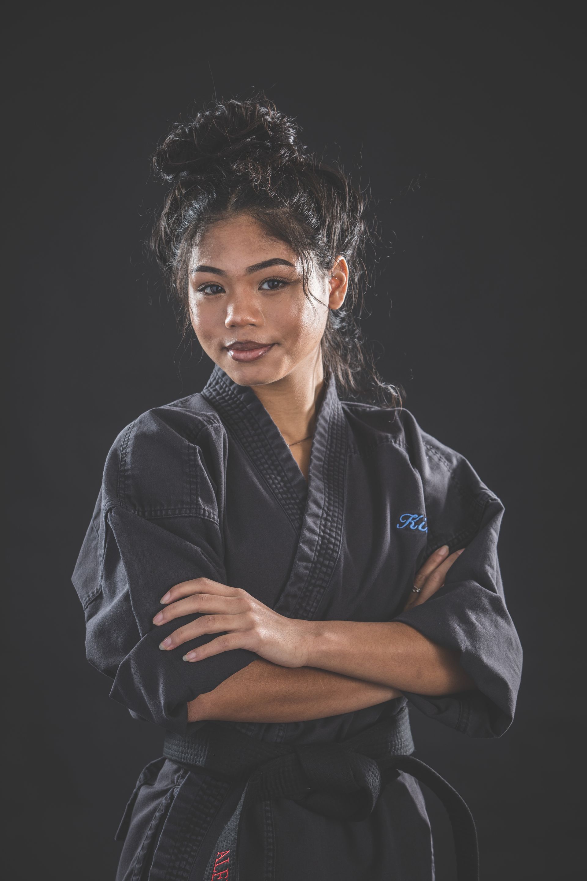 Person in a black martial arts uniform with arms crossed, standing against a dark background.