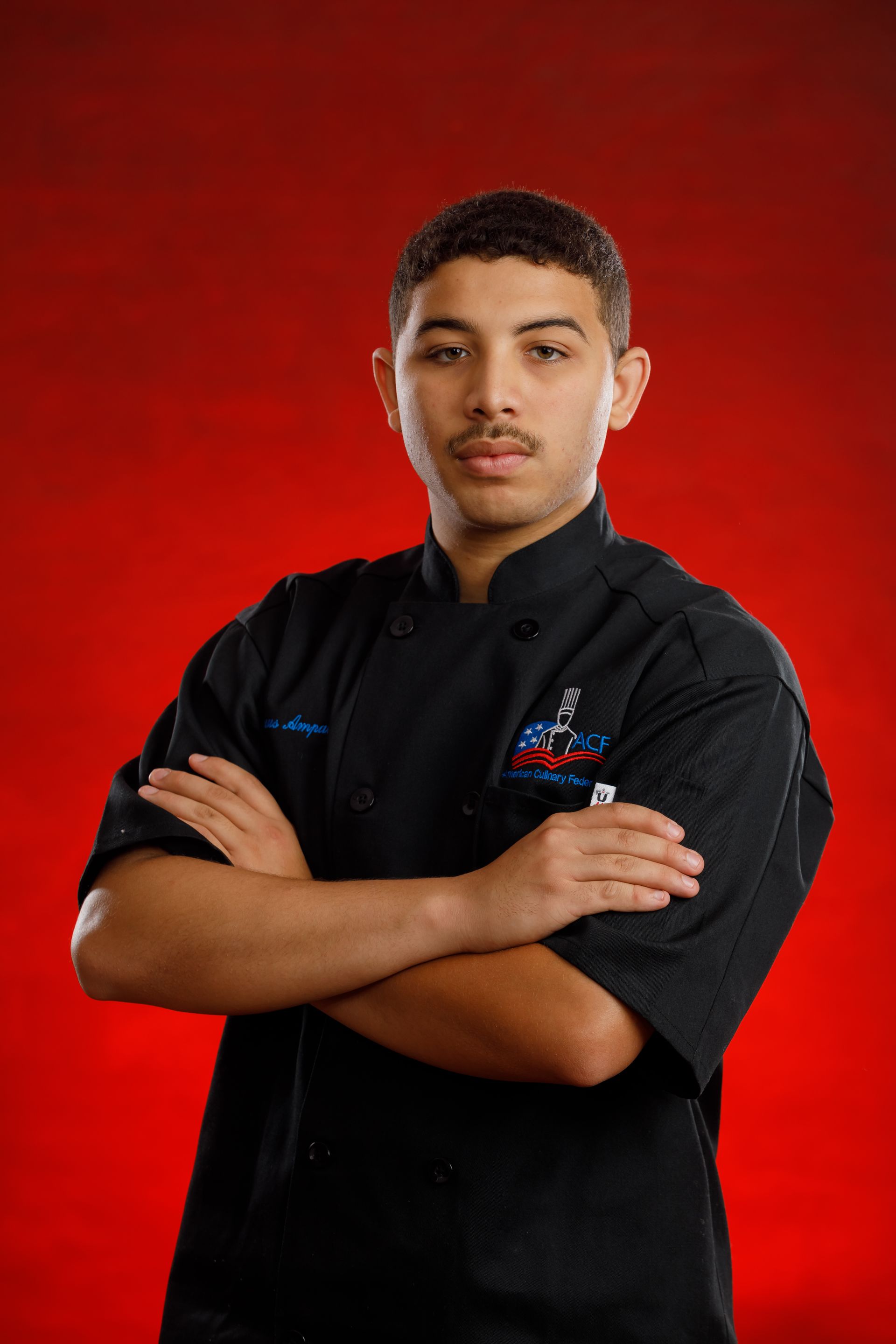 Person in a black chef coat with arms crossed against a red studio background