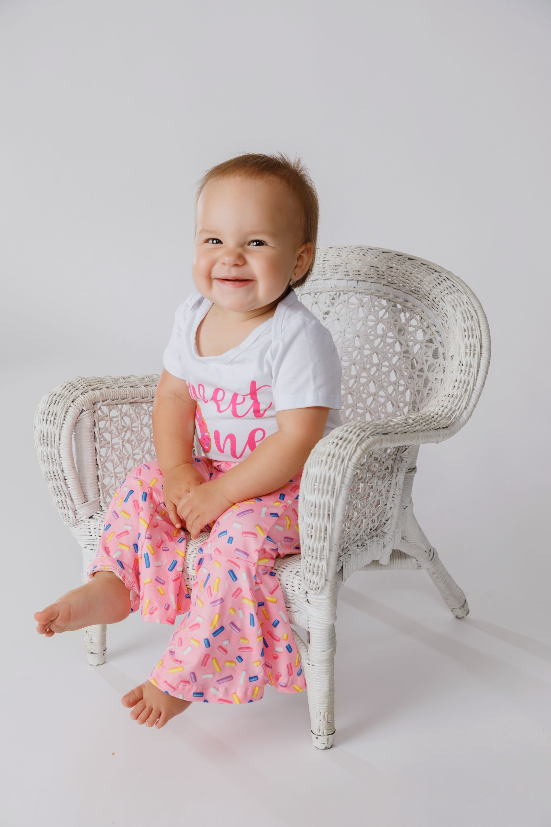 Smiling child sitting on a white wicker chair, wearing a white shirt and pink patterned skirt.