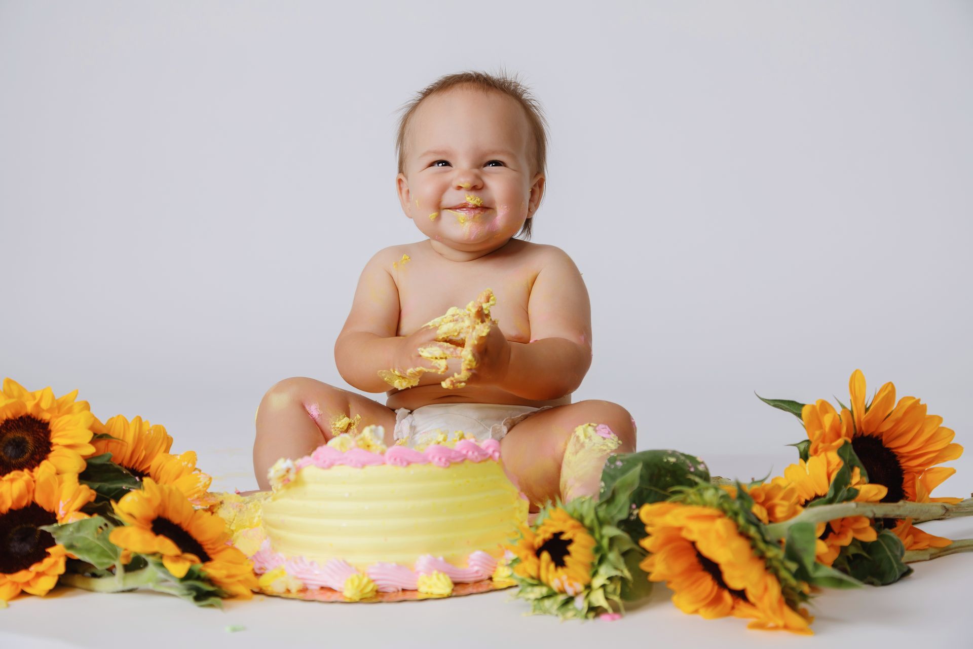Baby sitting on a pink cake surrounded by sunflowers, smiling in a bright studio setting
