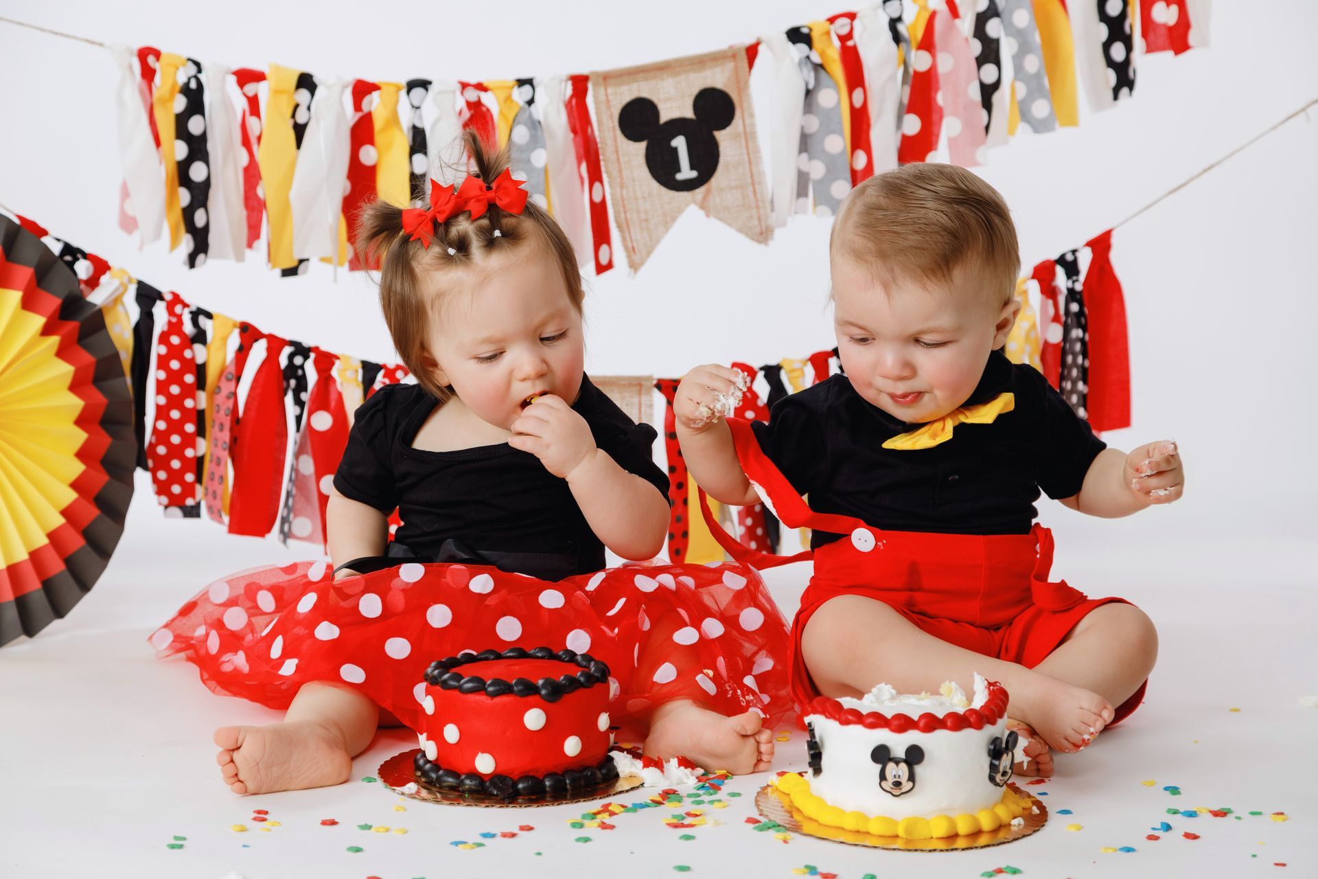 Two toddlers in red-and-black outfits eating cake at a Mickey Mouse-themed birthday party