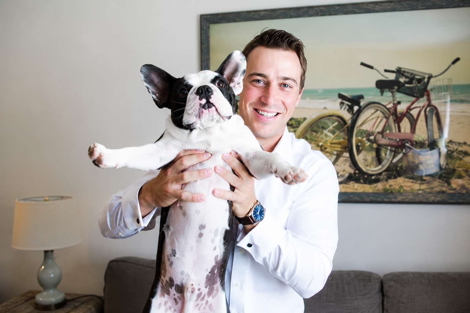 Man holding a black-and-white dog indoors, with a bicycle painting and lamp in the background