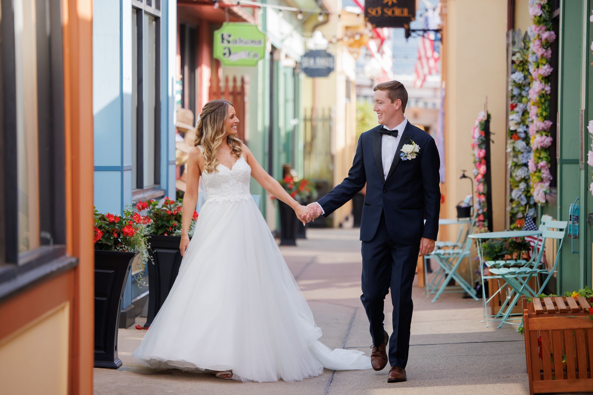 Bride and groom holding hands on a colorful street, walking past shops and flowers.