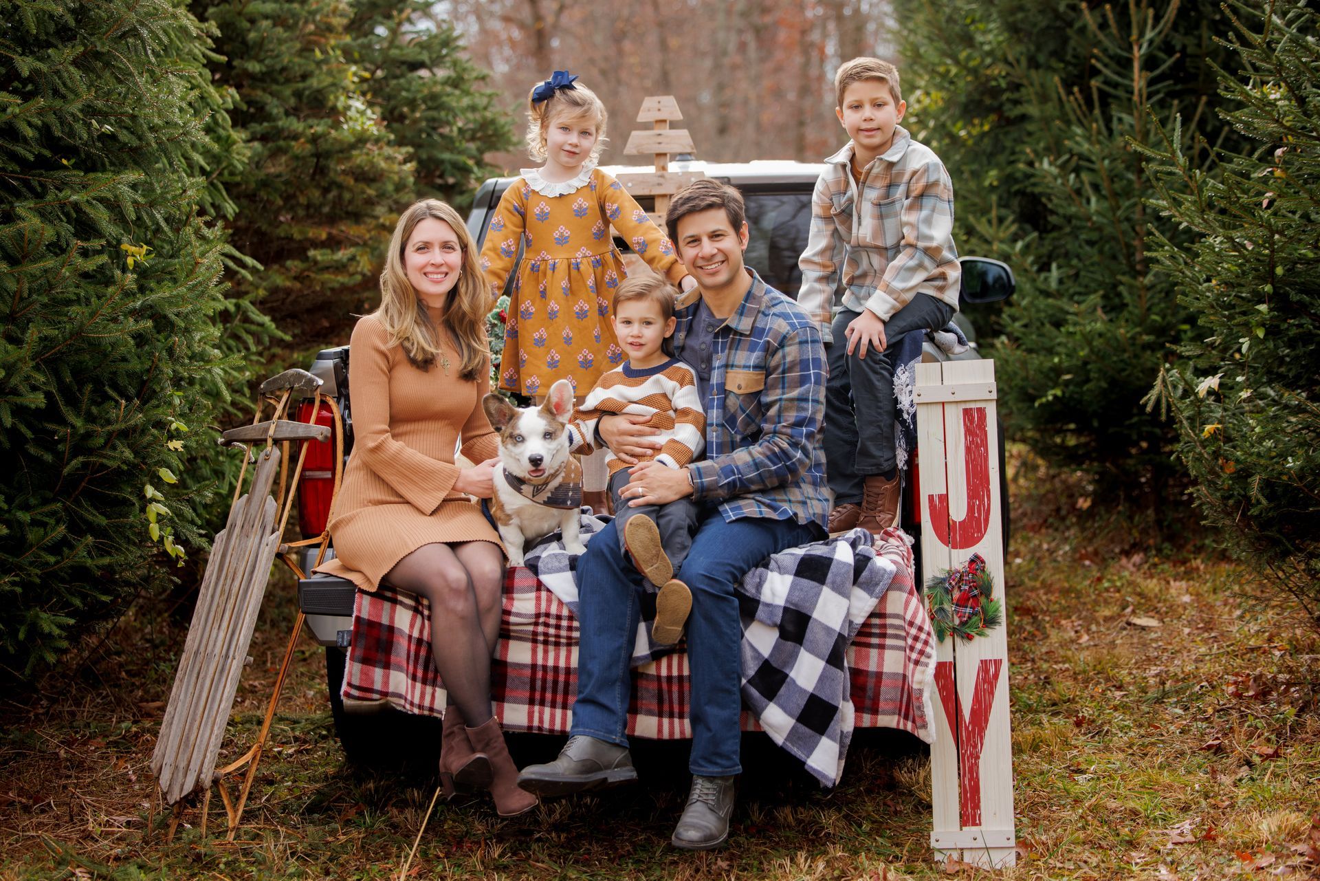 Family portrait on a plaid bench in a wooded setting, with a dog and a holiday sign reading “JOY”