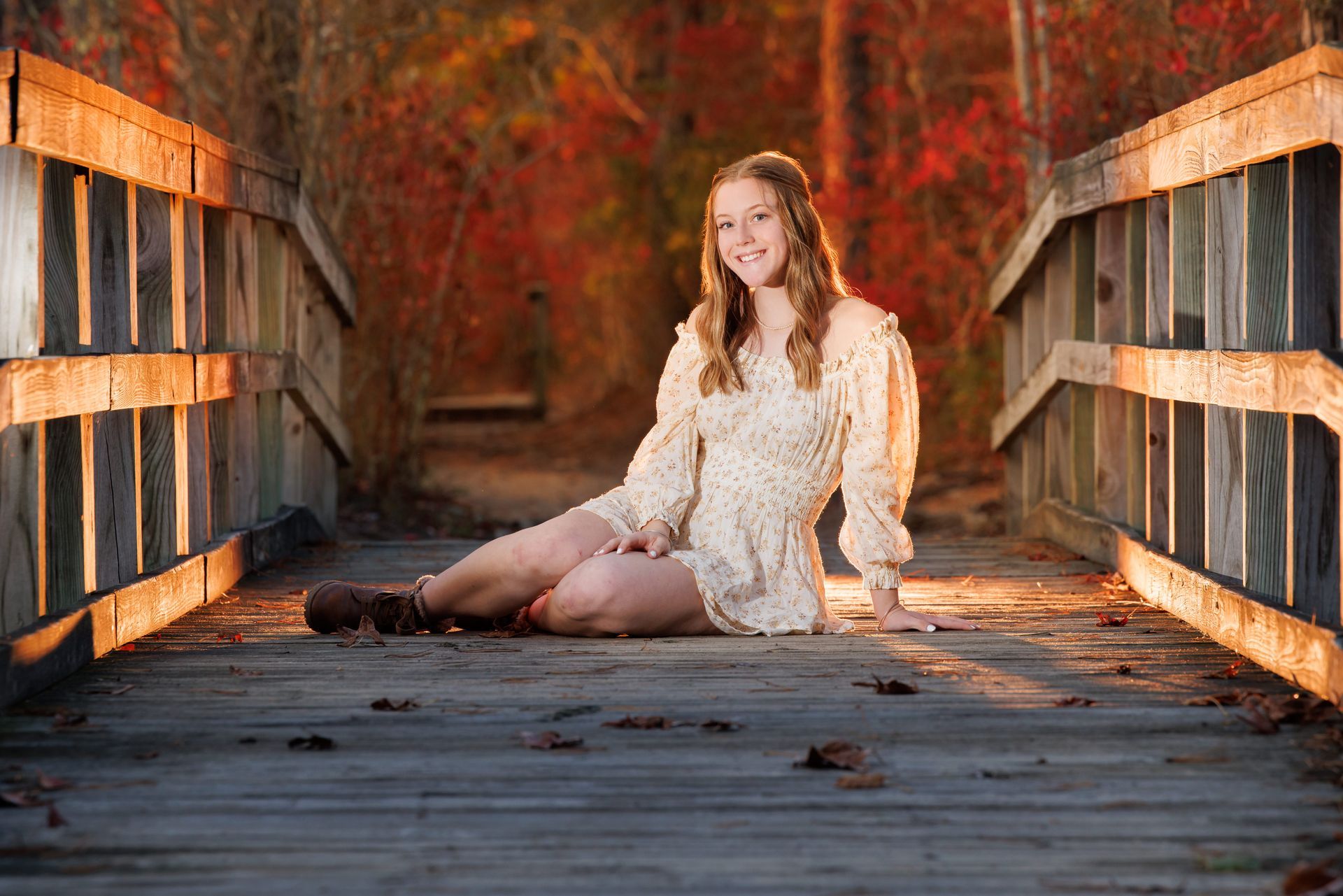 Woman in a white dress sitting on a wooden bridge, surrounded by warm autumn foliage.