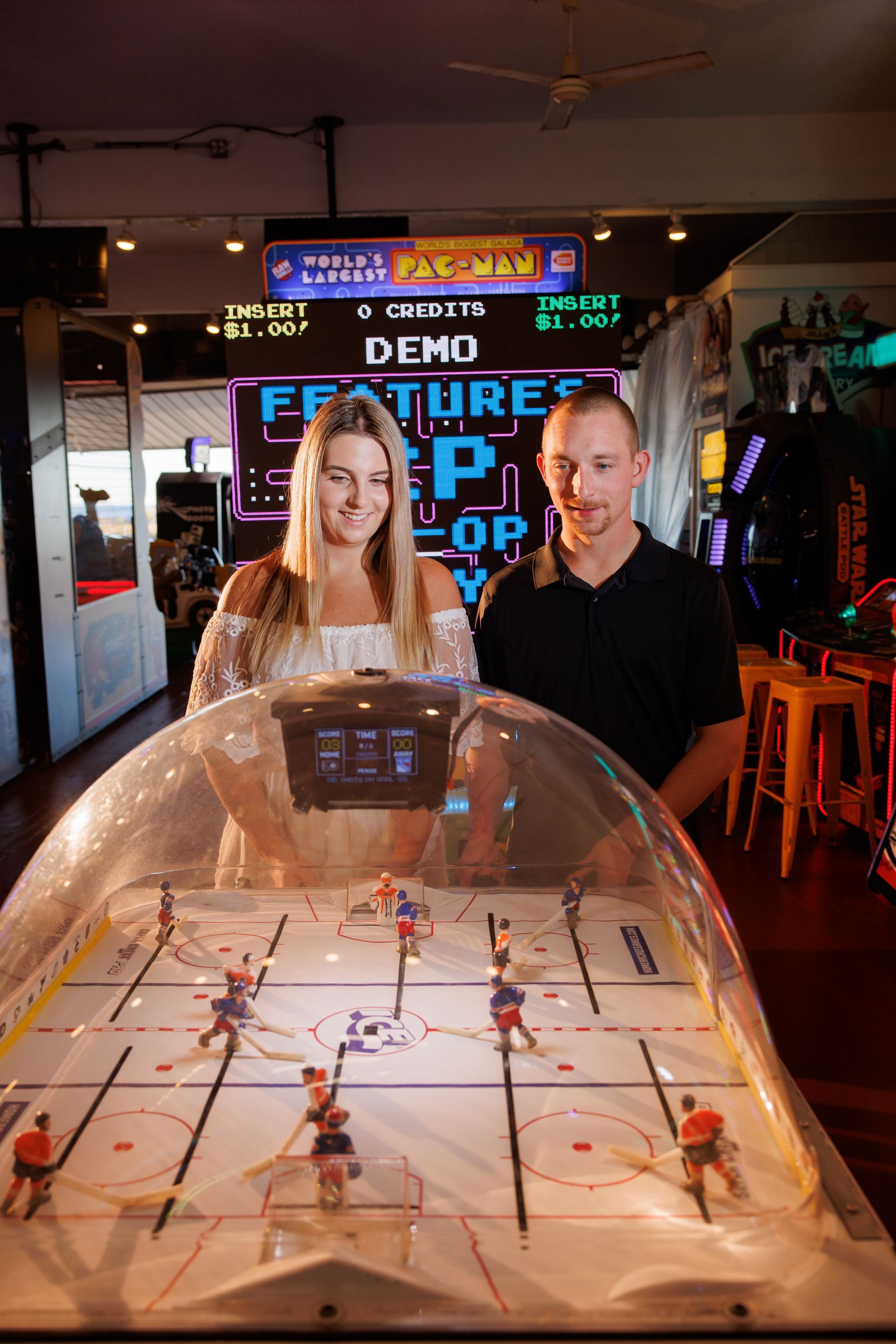 Two people stand behind a transparent arcade table game in a neon-lit arcade.