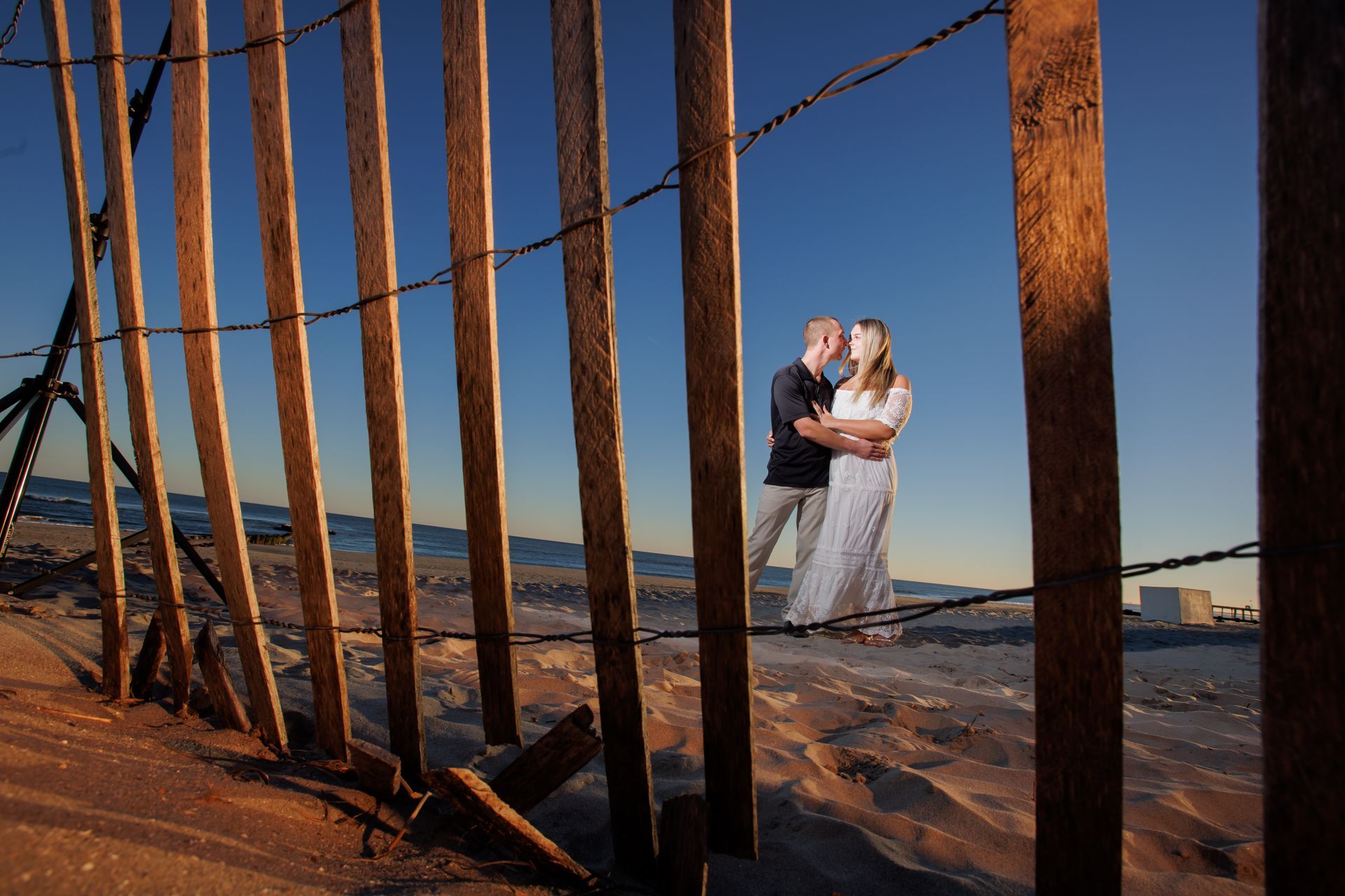 Couple embracing on a beach at sunset beside a row of wooden posts and a pier