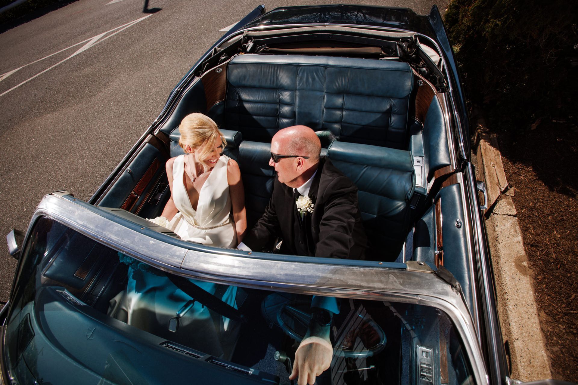 Two people in a black convertible, one holding a bouquet, viewed from above on a road
