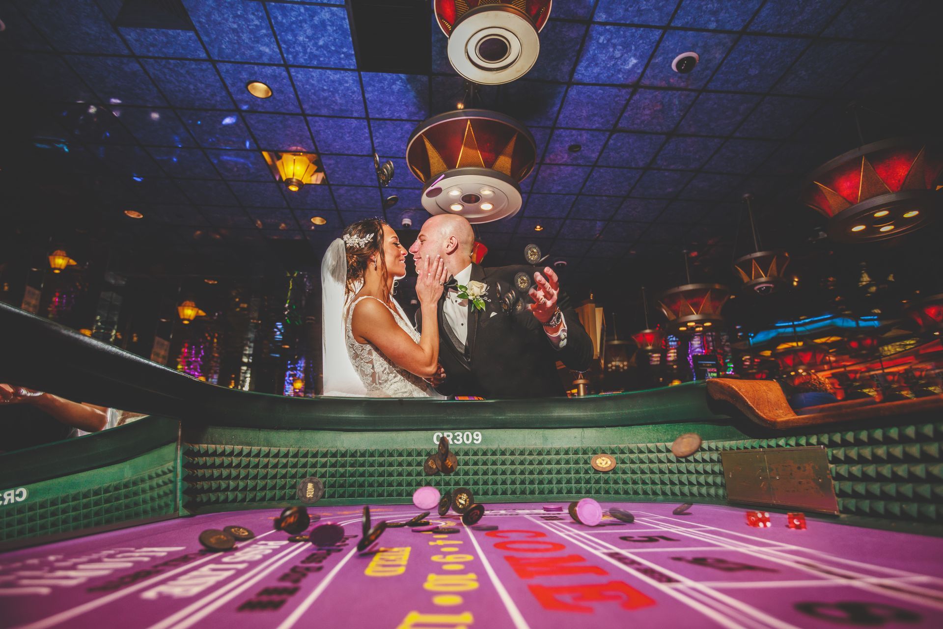 Bride and groom pose above a pink casino craps table under colorful ceiling lights
