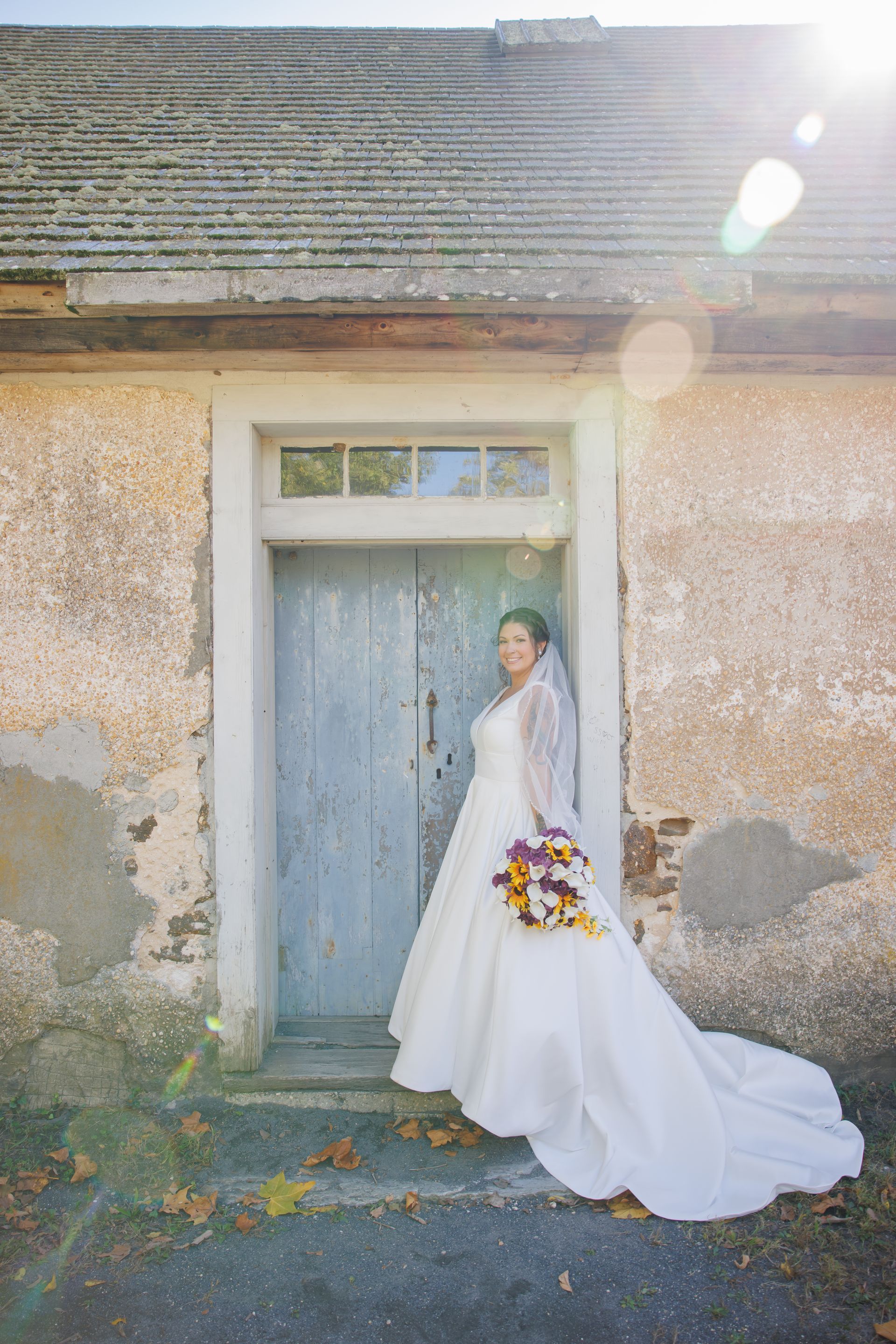Bride in a white gown holding a colorful bouquet, standing in a sunlit doorway of a rustic stone building