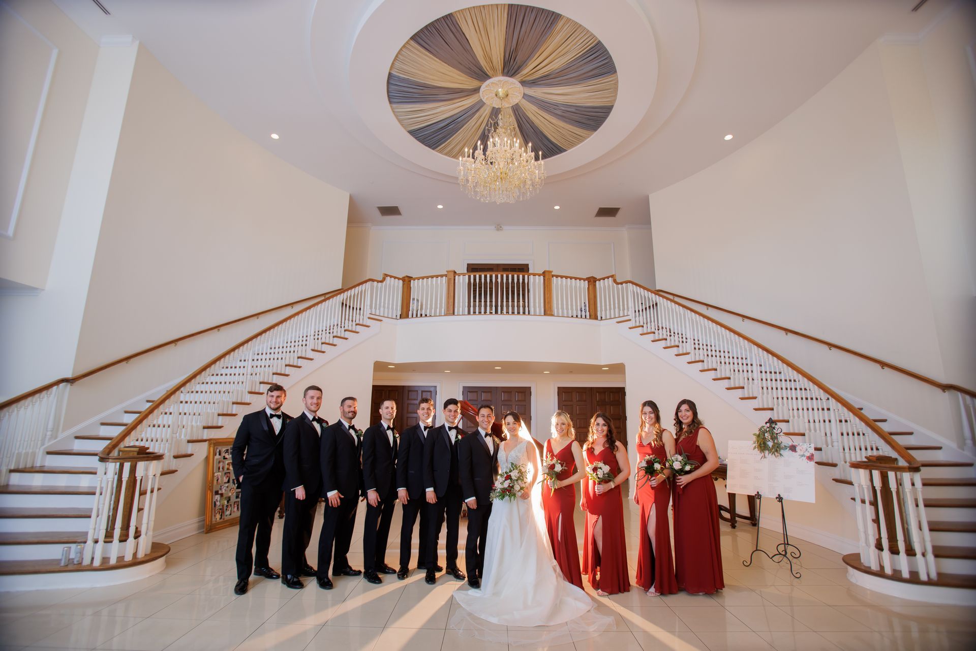 Wedding party posing in a grand white ballroom with a chandelier and sweeping staircase