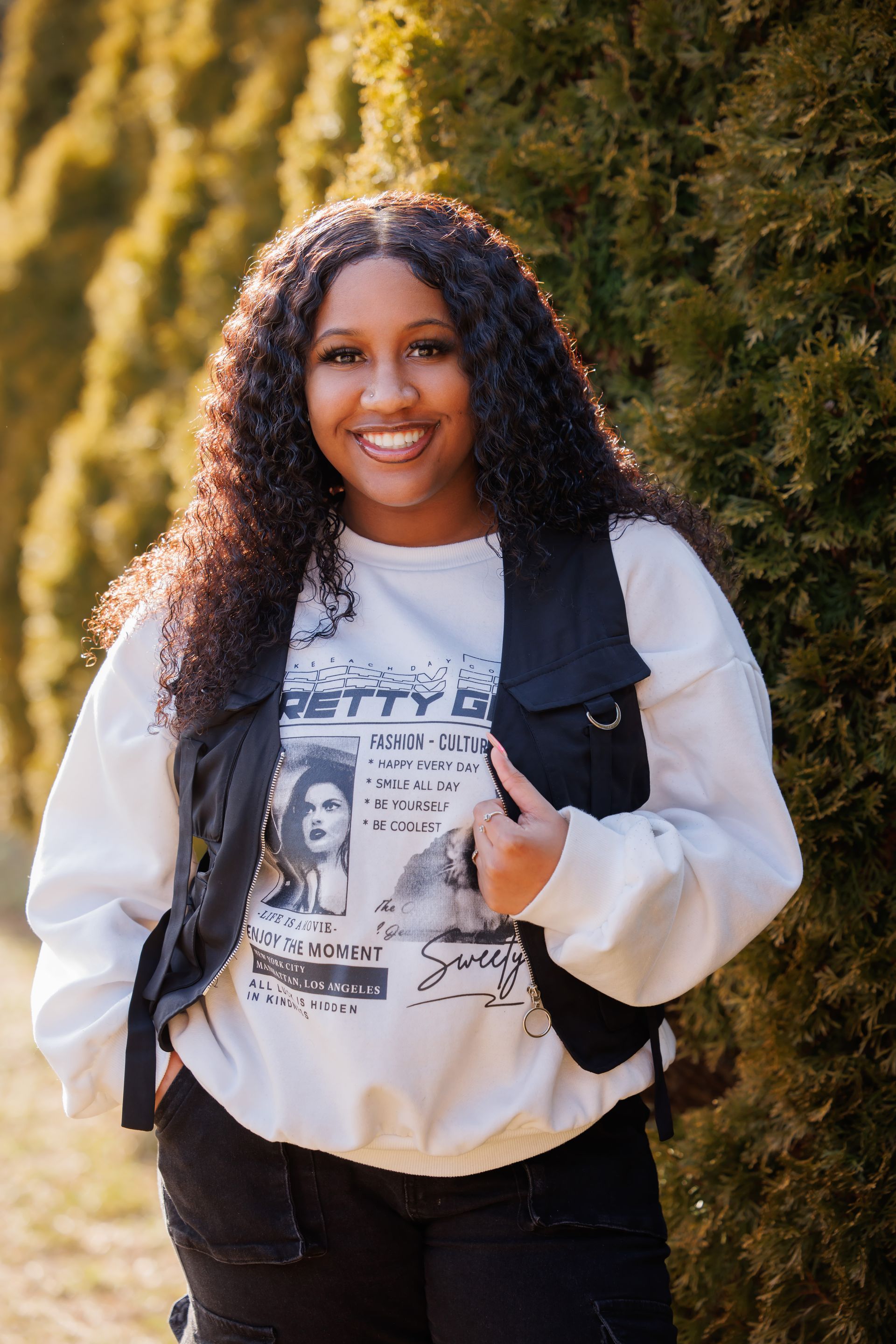 Smiling person in white sweatshirt and black vest standing outdoors on a sunlit path.