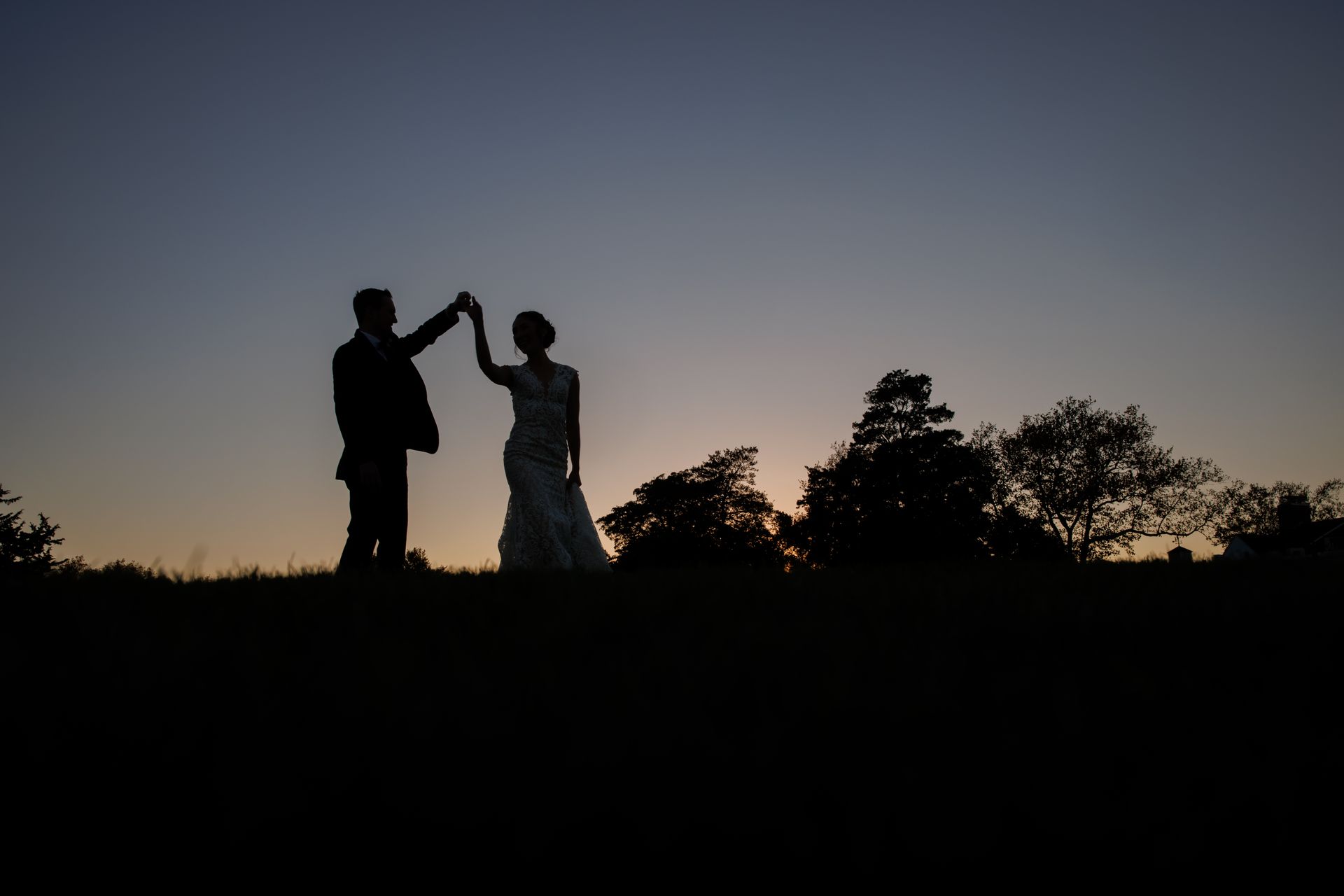 Silhouetted couple dancing at sunset on a grassy hill, with trees against the darkening sky.