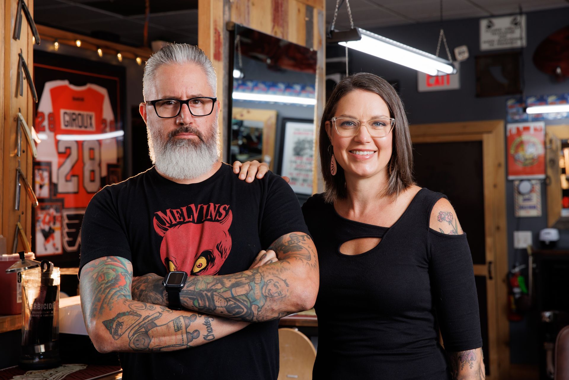 Two tattooed people standing in a bar, one with crossed arms, both smiling.