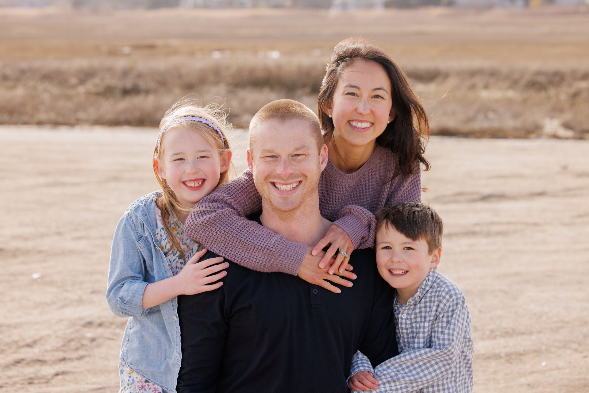 Three smiling children pose outdoors in a dry field, with two hugging a man in the center.