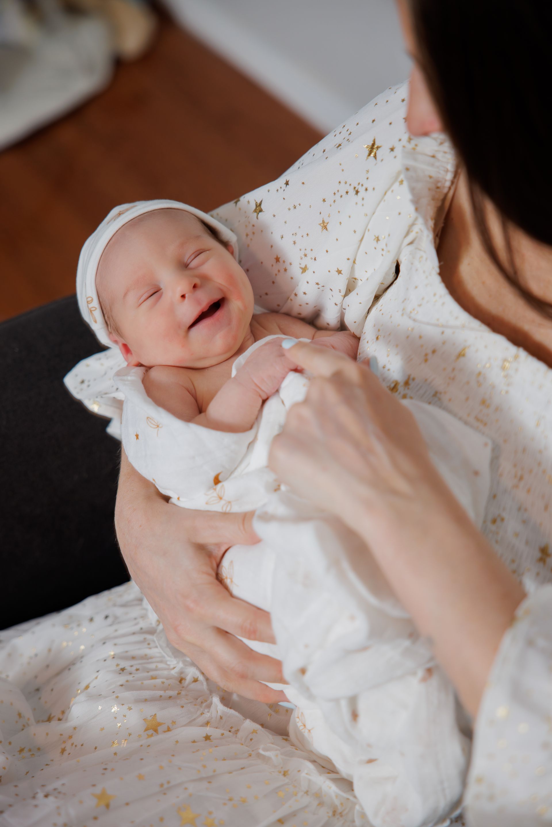 Smiling newborn wrapped in a white blanket, held by an adult in a cozy indoor setting.