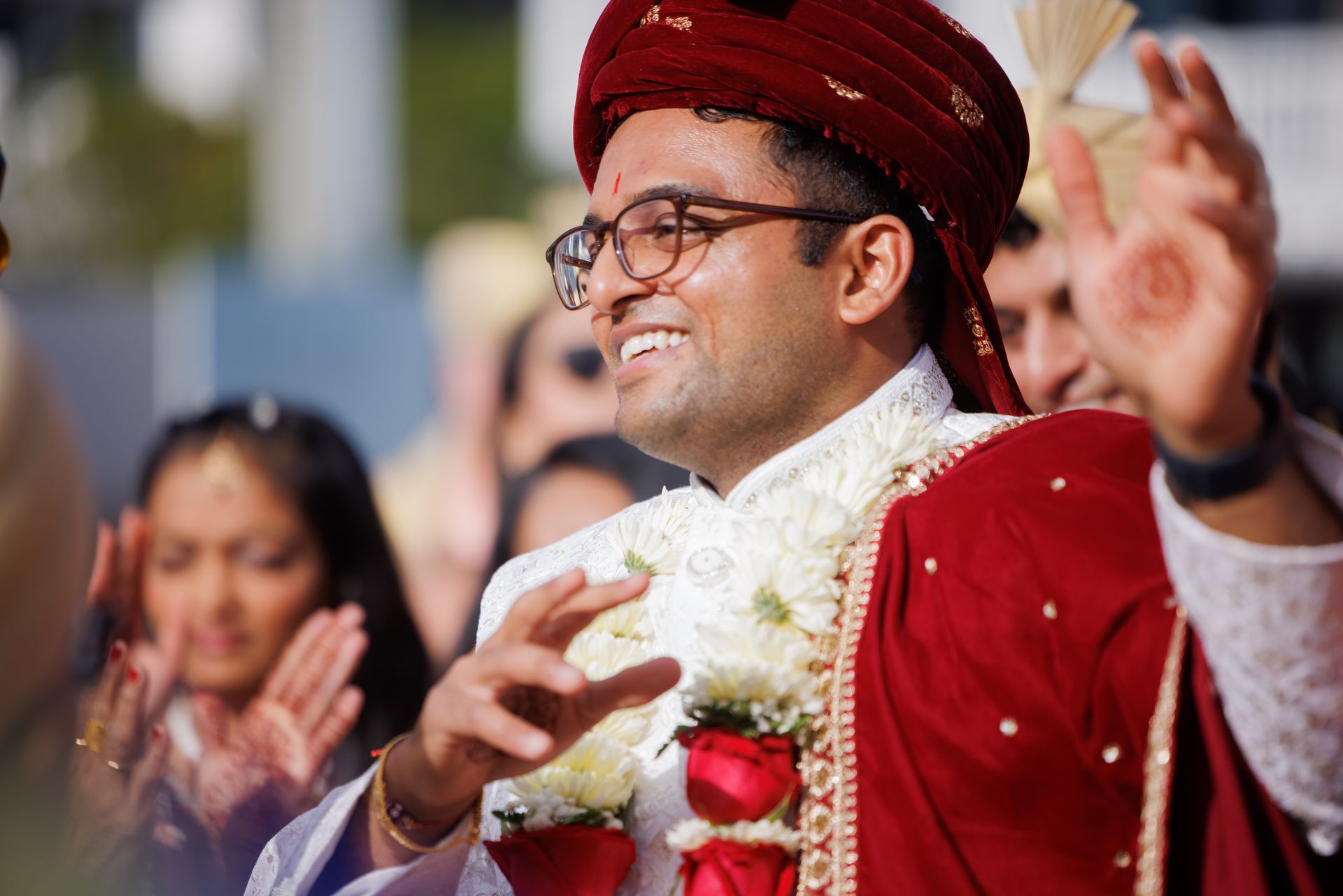 Smiling groom in red traditional attire with white garland, clapping at a wedding ceremony