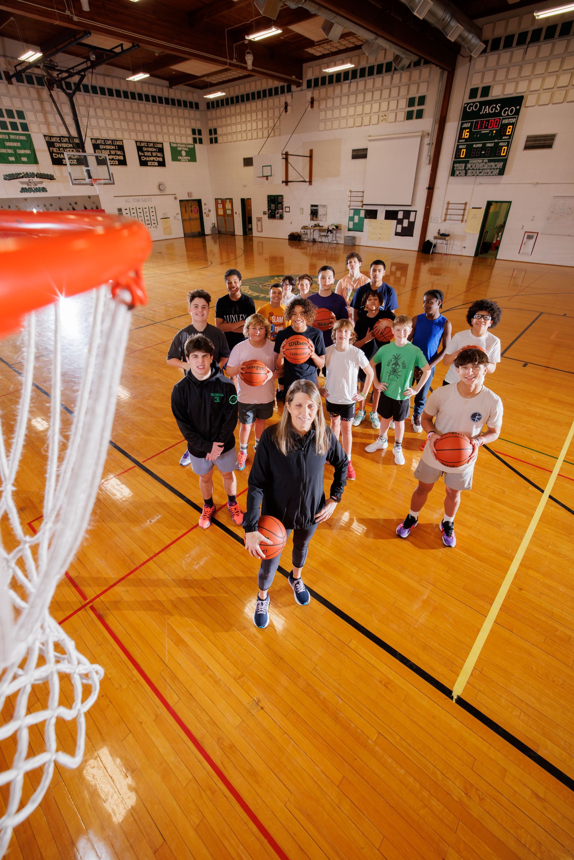 Children lined up on a gym floor near a basketball hoop, holding basketballs in a school gym