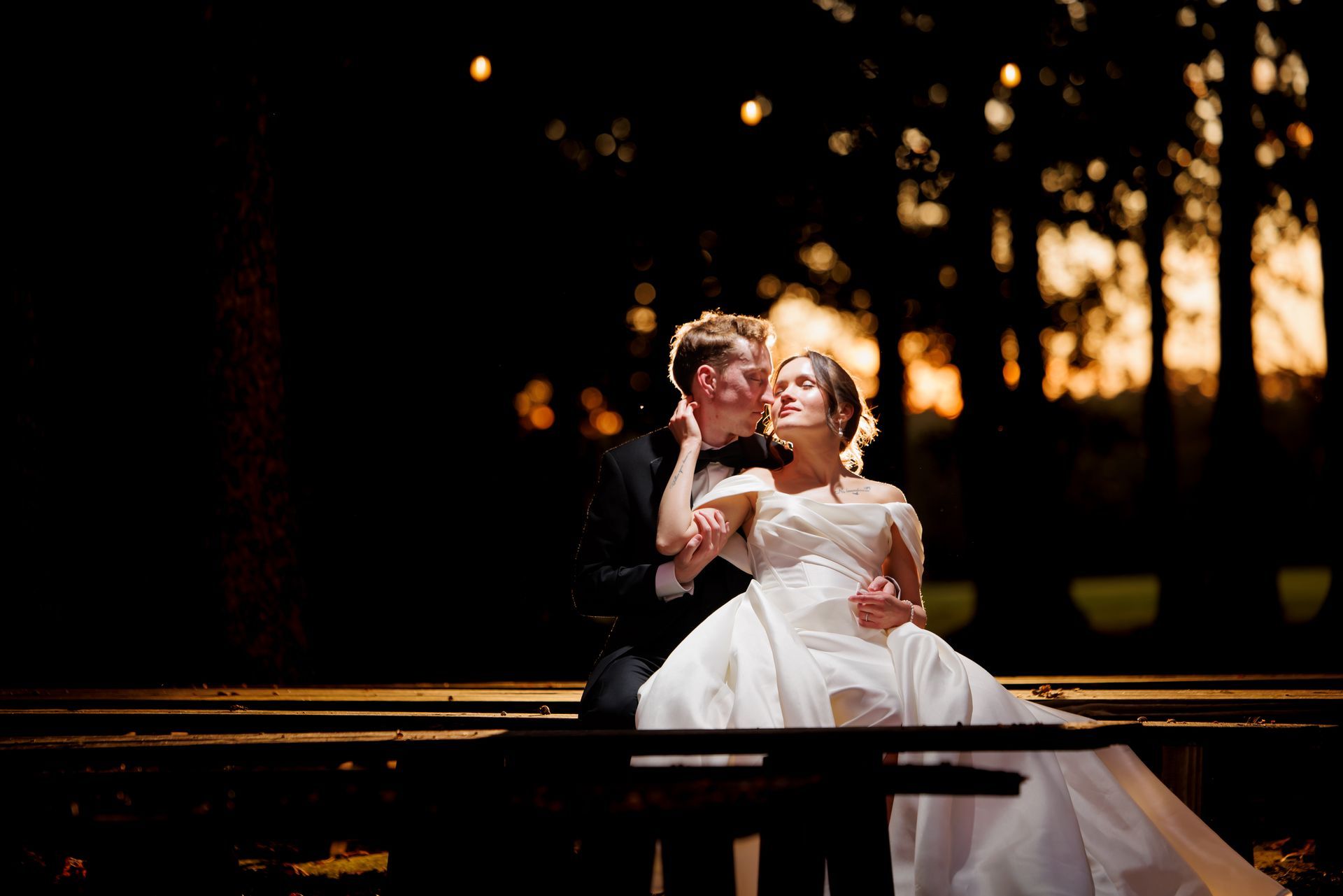 Wedding couple seated on a bench at sunset, sharing a tender moment under warm lights