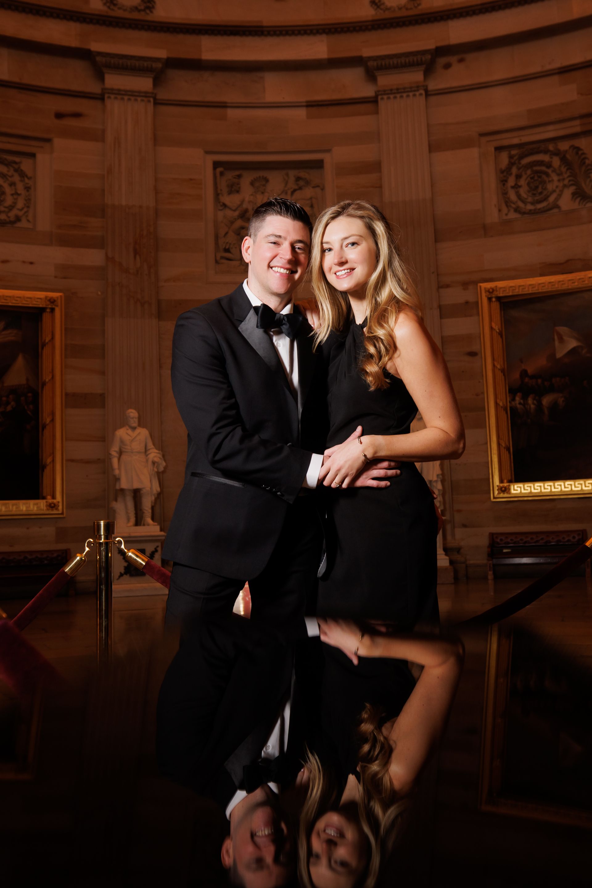 Couple in formal black attire posing in a grand hall with their reflection on a polished floor