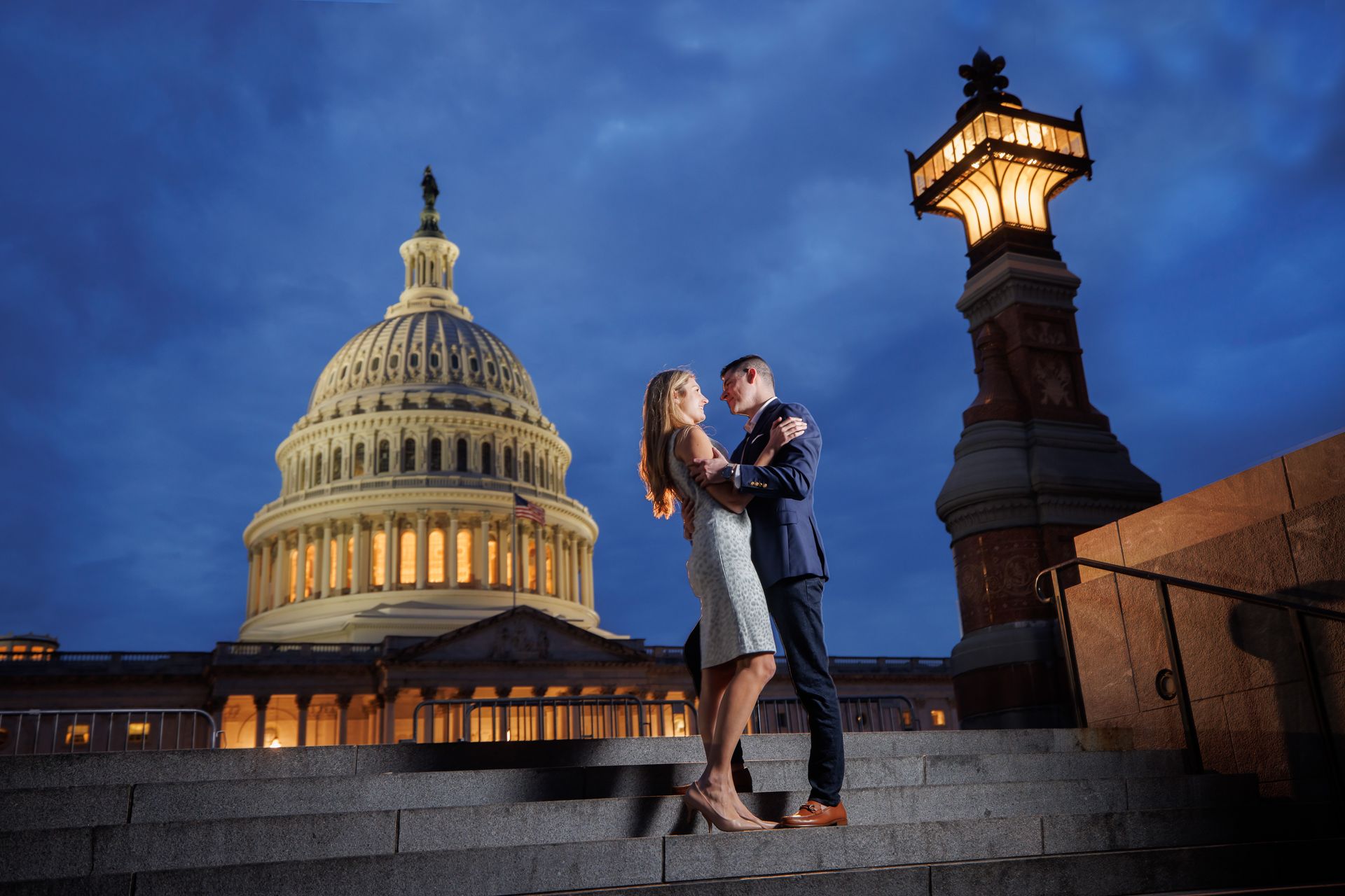 Couple embracing on the U.S. Capitol steps at dusk, with the lit Capitol dome and a glowing lamppost nearby