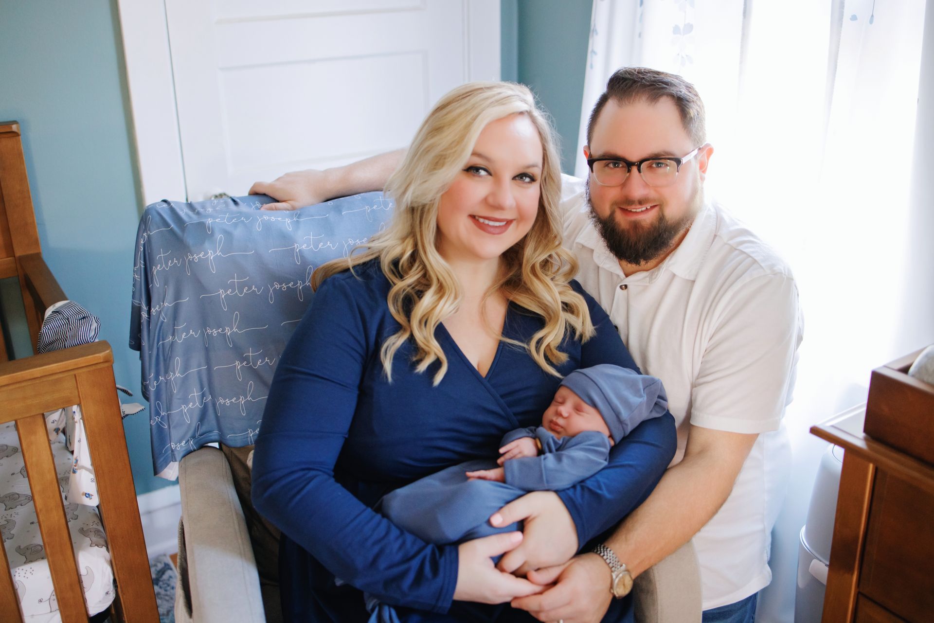 Couple holding a sleeping newborn in a bright nursery with a blue blanket and white crib railings
