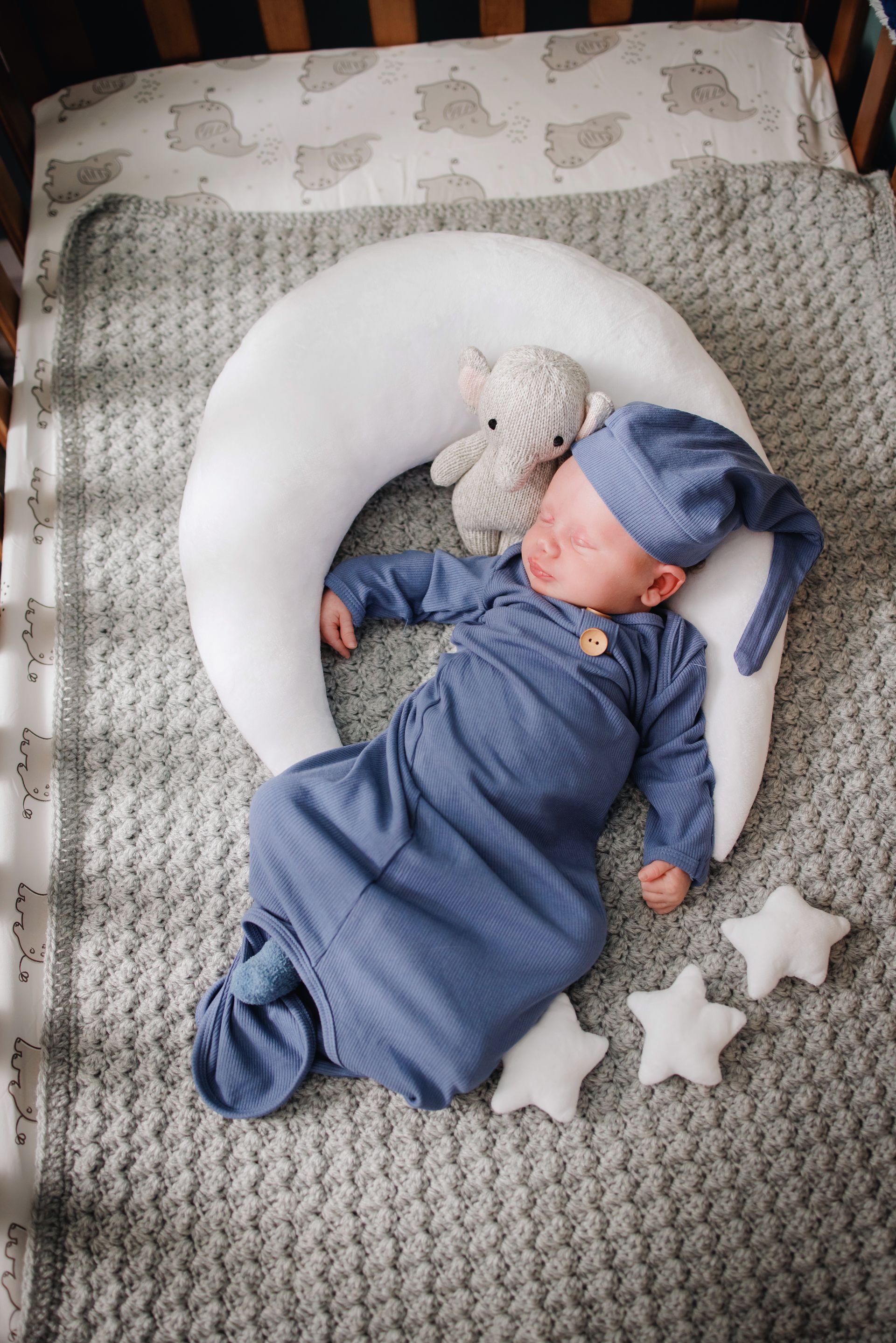 Sleeping baby in blue pajamas on a crescent moon pillow, with a stuffed toy and star cushions on a gray blanket.