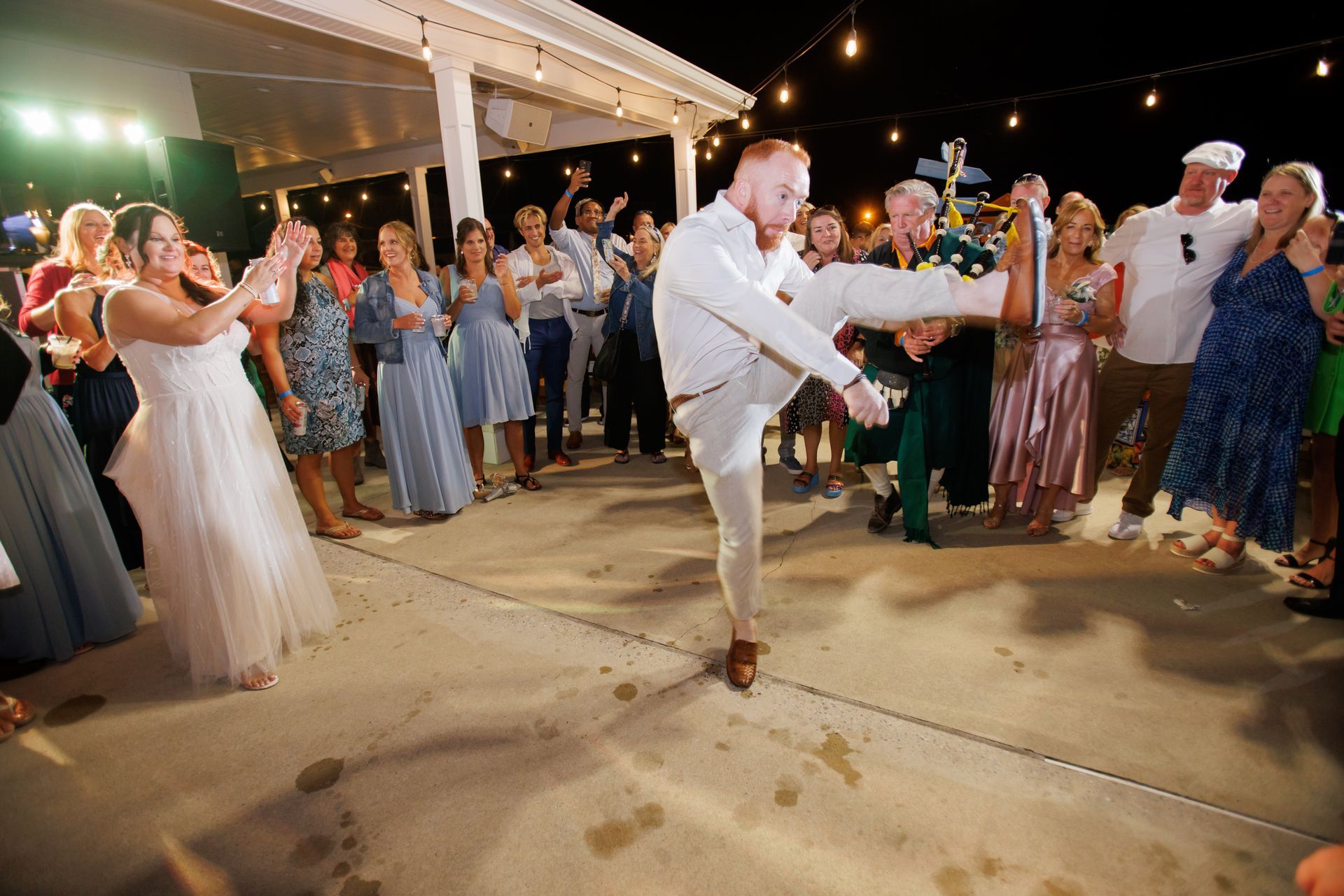Bride and groom dance at an outdoor wedding reception as guests clap and cheer around them.