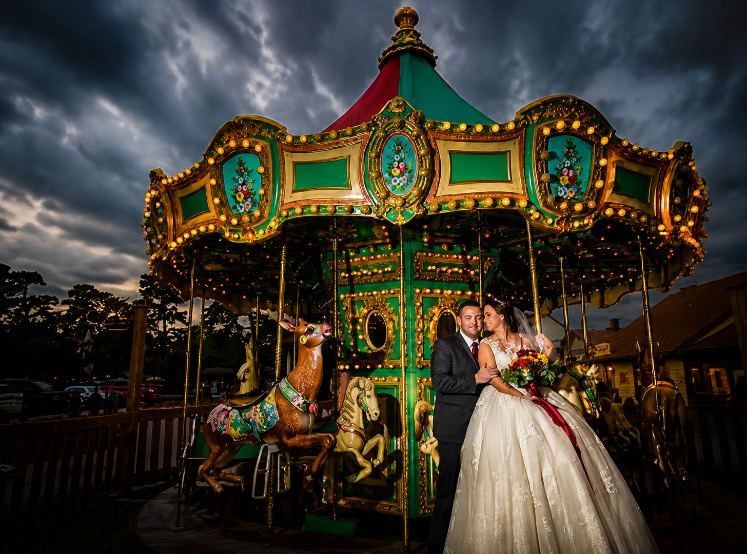 Bride in white dress on a colorful carousel horse at night, with a lit vintage carousel behind her