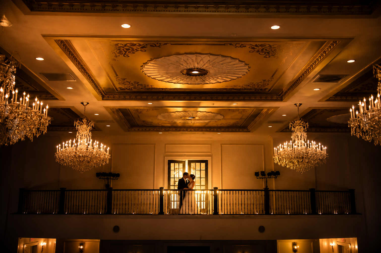 Elegant ballroom ceiling with ornate molding and crystal chandeliers over a balcony.