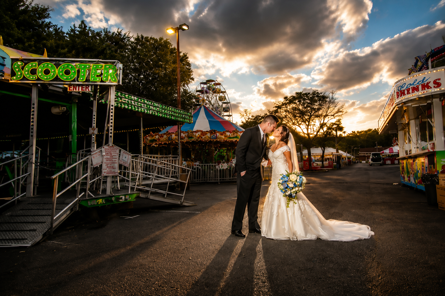 Bride and groom kissing in a carnival midway under a cloudy sky