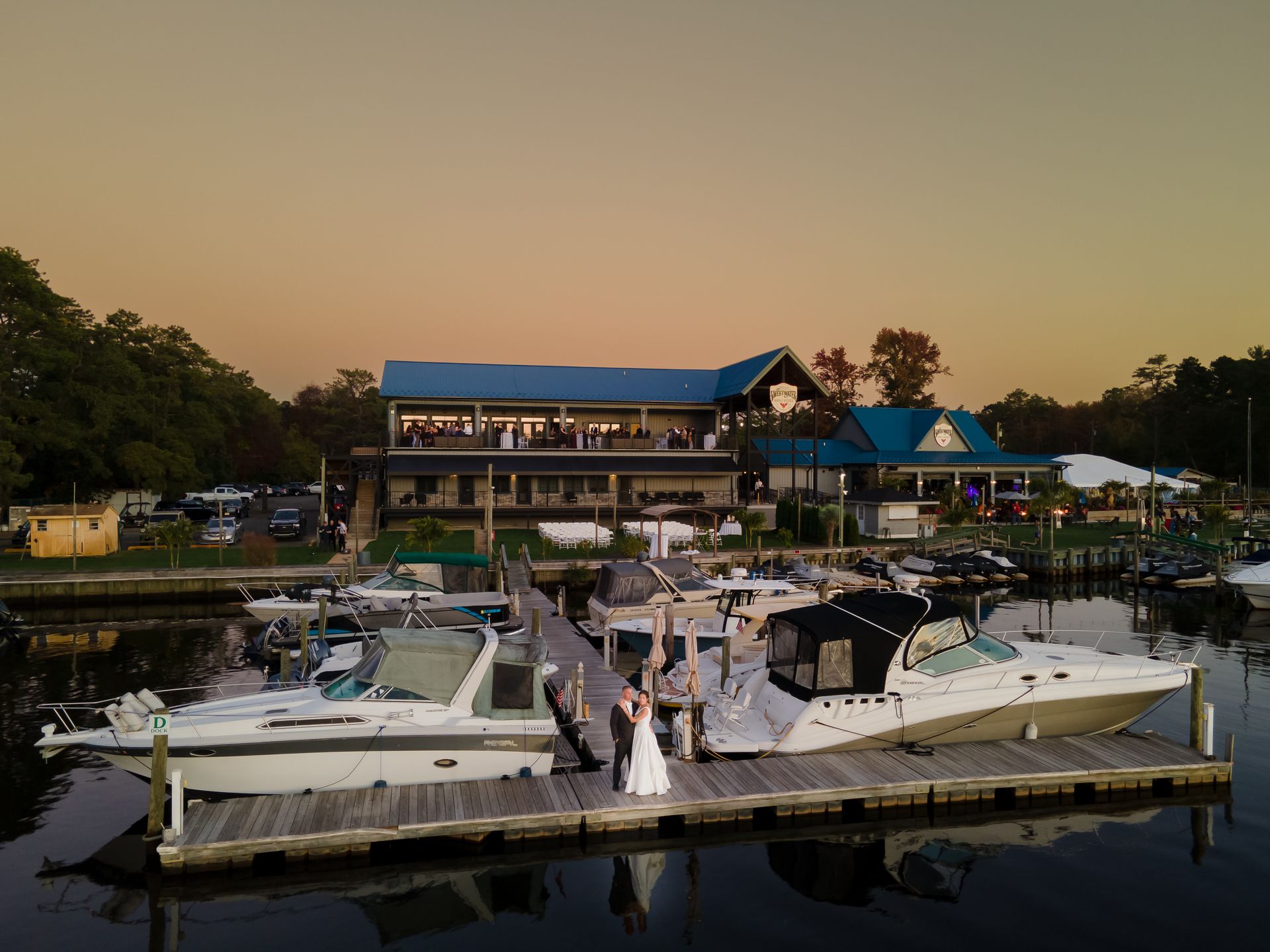 Yachts docked at a marina with people on the pier at sunset, buildings and trees in the background.