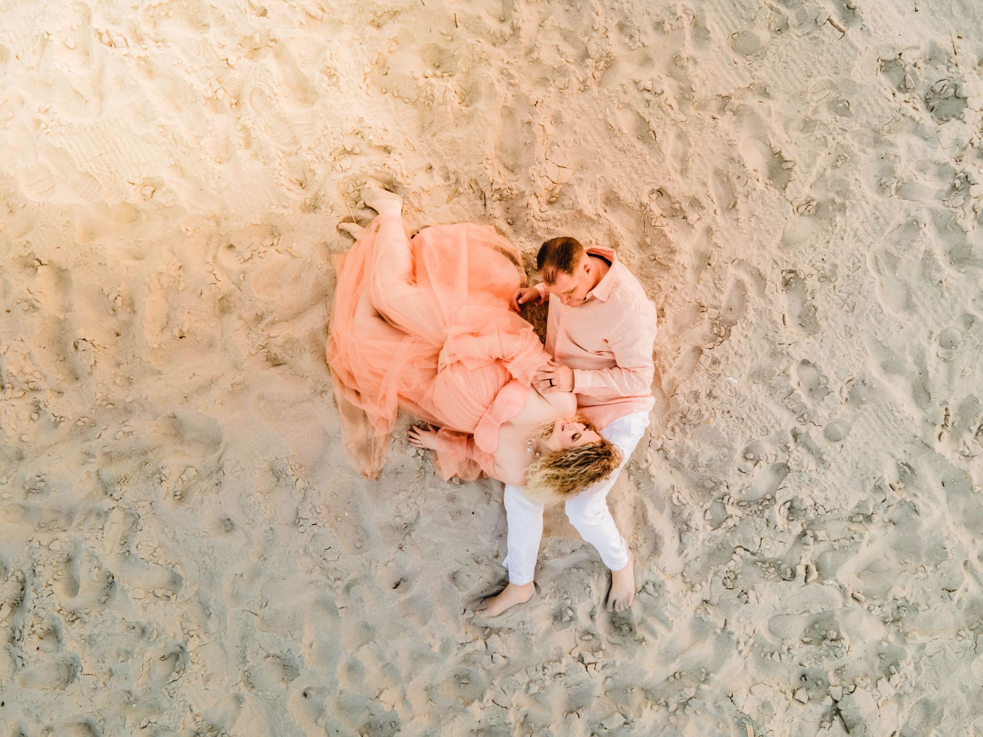 Couple lying on sandy beach, wrapped together in a soft pink blanket.